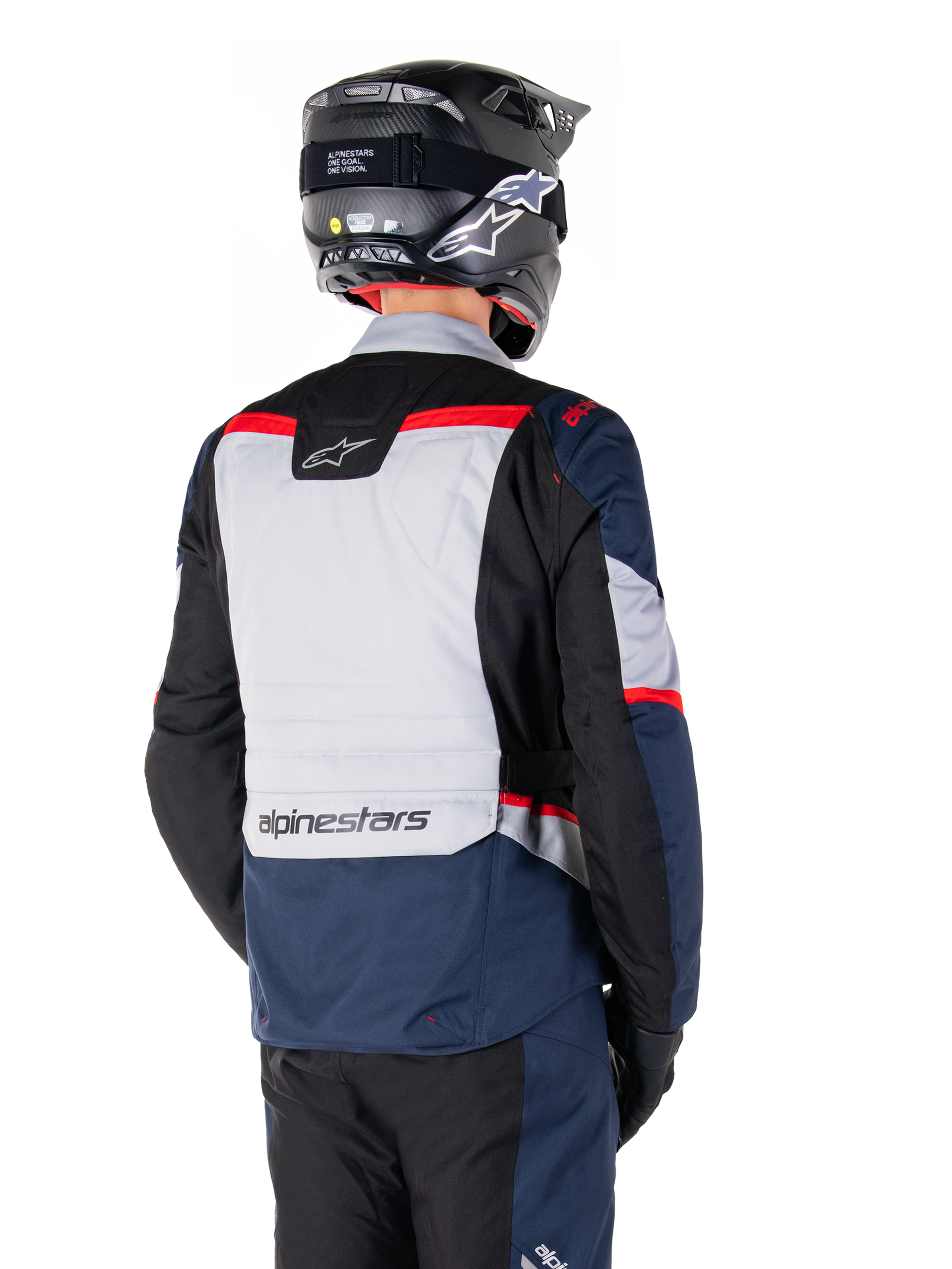 Chaqueta impermeable Alpinestars ST-1, azul oscuro, negro y rojo brillante, vista trasera de un piloto con casco de aventura negro, paneles en gris claro y marca Alpinestars, equipamiento de moto para turismo de aventura
