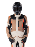 Chaqueta impermeable Alpinestars ST-1, Dark Khaki Sand Black Red Fluo, usada por un piloto con casco y guantes negros, equipamiento de touring de aventura con paneles beige y marrones y detalles en rojo flúor brillante, vista frontal sobre fondo blanco.