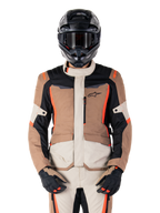 Chaqueta impermeable Alpinestars ST-1, Dark Khaki Sand Black Red Fluo, usada por un piloto con casco y guantes negros, equipamiento de touring de aventura con paneles beige y marrones y detalles en rojo flúor brillante, vista frontal sobre fondo blanco.