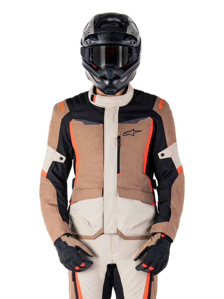 Chaqueta impermeable Alpinestars ST-1, Dark Khaki Sand Black Red Fluo, usada por un piloto con casco y guantes negros, equipamiento de touring de aventura con paneles beige y marrones y detalles en rojo flúor brillante, vista frontal sobre fondo blanco.