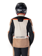 Chaqueta impermeable Alpinestars ST-1, Dark Khaki Sand Black Red Fluo, vista trasera, chaqueta de motociclismo adventure touring con un gran panel trasero de color arena, mangas negras y detalles en rojo fluorescente, mostrada en un piloto con casco negro.