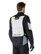 Alpinestars ST-1 Waterproof Jacket in Ice Gray Black Yellow Fluo, rear three-quarter view of a rider wearing the adventure touring jacket, featuring a large gray back panel with fluorescent yellow accents and Alpinestars branding, paired with black riding pants and gloves