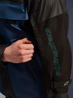 Alpinestars Mospher Waterproof Jacket in Black Dark Petrol, close-up detail of the sleeve with teal Alpinestars branding, a hand adjusting the ventilation zipper, featuring durable ripstop fabric and an adjustable arm strap