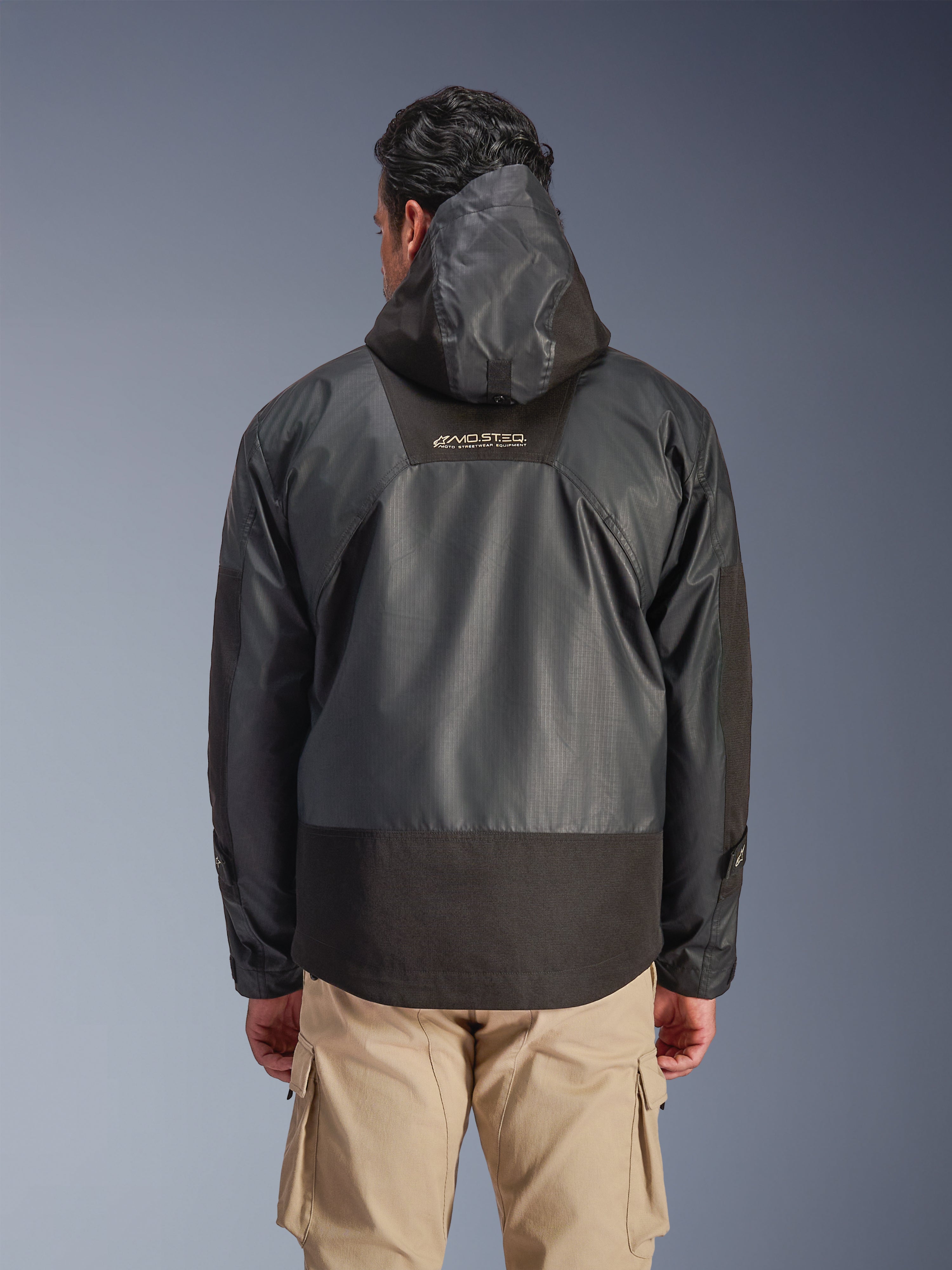 Vista posterior de un hombre con la chaqueta impermeable Alpinestars Mospher en color negro, con capucha y confección en tejido ripstop duradero, mostrada con pantalones cargo color tostado, destacando el logotipo MO.ST.EQ. en la parte superior de la espalda, diseño de equipamiento urbano para motociclista.