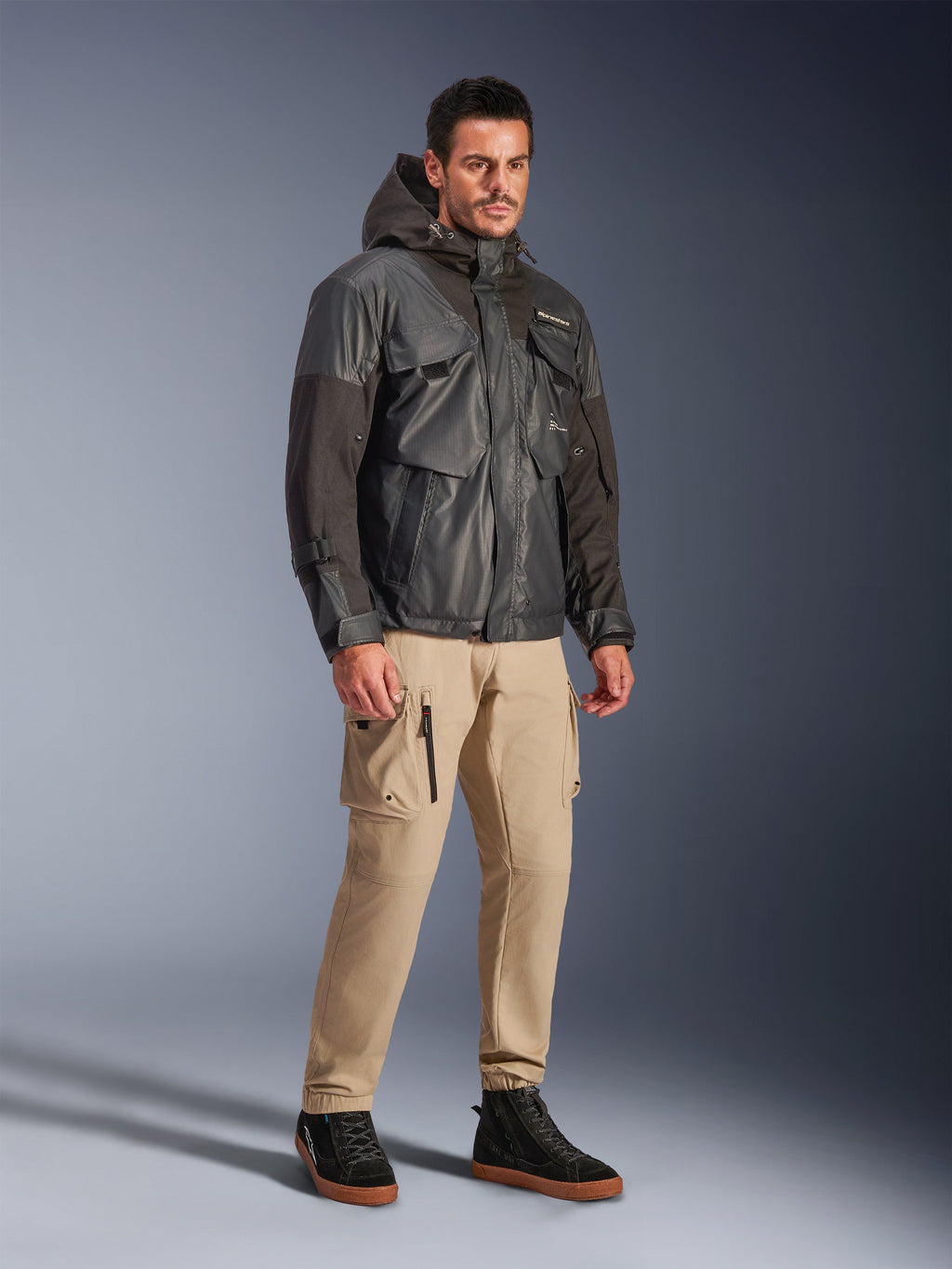 Modelo masculino vistiendo la chaqueta impermeable Alpinestars Mospher en color negro, parka impermeable de inspiración utilitaria con diseño multibolsillos y capucha, combinada con pantalones cargo color canela y zapatillas altas negras, equipamiento urbano para motociclistas.