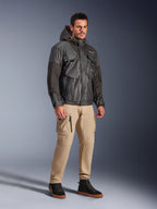 Modelo masculino vistiendo la chaqueta impermeable Alpinestars Mospher en color negro, parka impermeable de inspiración utilitaria con diseño multibolsillos y capucha, combinada con pantalones cargo color canela y zapatillas altas negras, equipamiento urbano para motociclistas.