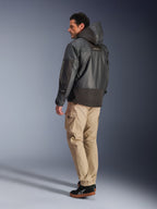 Hombre vistiendo la chaqueta impermeable Alpinestars Mospher en color negro, vista de perfil lateral y trasera, combinada con pantalones cargo color canela y zapatillas negras. Presenta un diseño de parka con capucha en tejido ripstop, marca Alpinestars en la manga y un estilo de motociclismo urbano moderno.