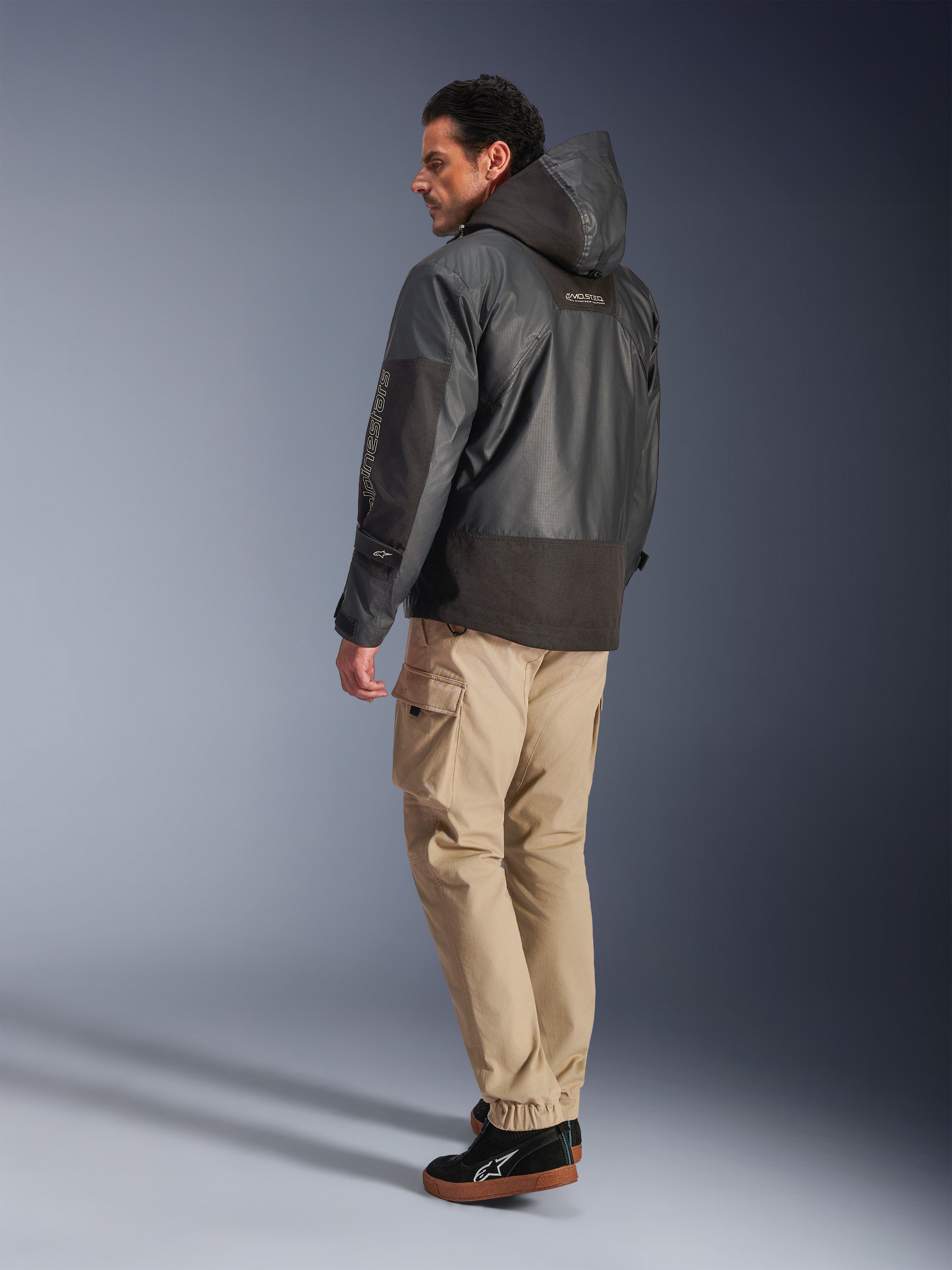 Hombre vistiendo la chaqueta impermeable Alpinestars Mospher en color negro, vista de perfil lateral y trasera, combinada con pantalones cargo color canela y zapatillas negras. Presenta un diseño de parka con capucha en tejido ripstop, marca Alpinestars en la manga y un estilo de motociclismo urbano moderno.