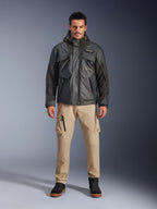 Modelo masculino luciendo la chaqueta impermeable Alpinestars Mospher en color negro, equipamiento de moto urbano contemporáneo, diseño táctico funcional con múltiples bolsillos y capucha, combinado con pantalones cargo color arena y calzado negro, retrato de estudio de cuerpo completo.