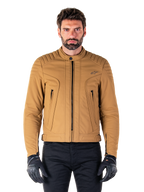 Chaqueta Alpinestars Clayton WR, chaqueta de moto urbana, color Utility Brown Light Gold, textil resistente al agua con hombros acolchados, cuello alto de motorista, detalles de cremalleras negras y logo en el pecho, usada con guantes de protección y pantalones negros