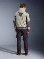 Chaqueta impermeable Alpinestars Moblast, Iron Gray Dark Sage, vista trasera de un piloto masculino con la chaqueta de moto con capucha, combinación de colores gris claro y verde salvia oscuro, logotipo 'astars' vertical en la parte superior de la espalda, parches gris oscuro en los codos, combinada con pantalones cargo oscuros para un look urbano contemporáneo.
