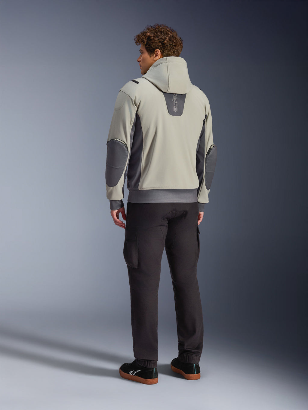 Modelo masculino visto desde atrás luciendo la chaqueta Alpinestars Moblast Waterproof en color Iron Gray Dark Sage, chaqueta urbana para moto con capucha con paneles laterales gris oscuro y parches reforzados en los codos, combinada con pantalones de conducción oscuros y zapatillas negras, estética minimalista moderna para desplazamientos urbanos.
