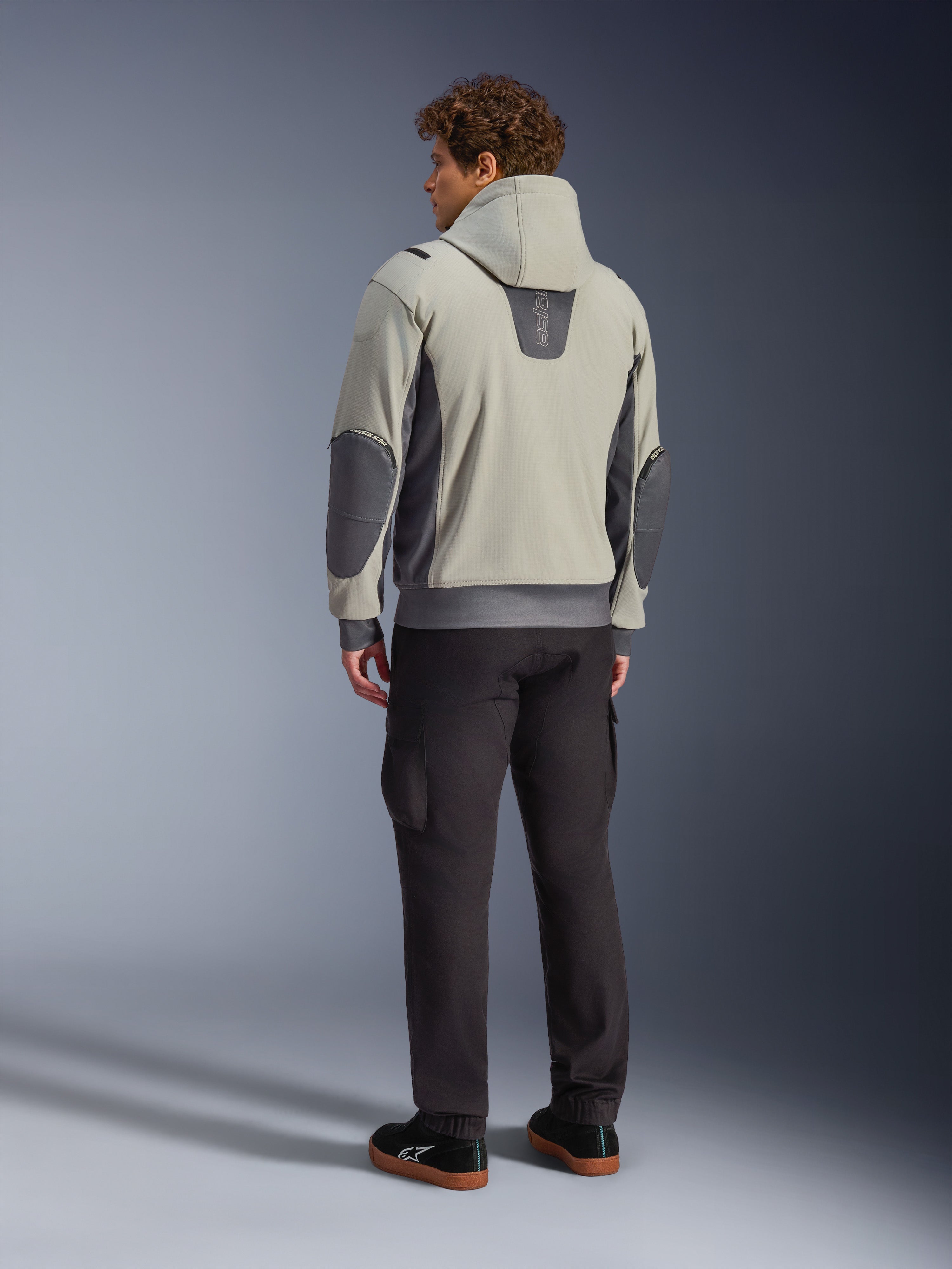 Modelo masculino visto desde atrás luciendo la chaqueta Alpinestars Moblast Waterproof en color Iron Gray Dark Sage, chaqueta urbana para moto con capucha con paneles laterales gris oscuro y parches reforzados en los codos, combinada con pantalones de conducción oscuros y zapatillas negras, estética minimalista moderna para desplazamientos urbanos.