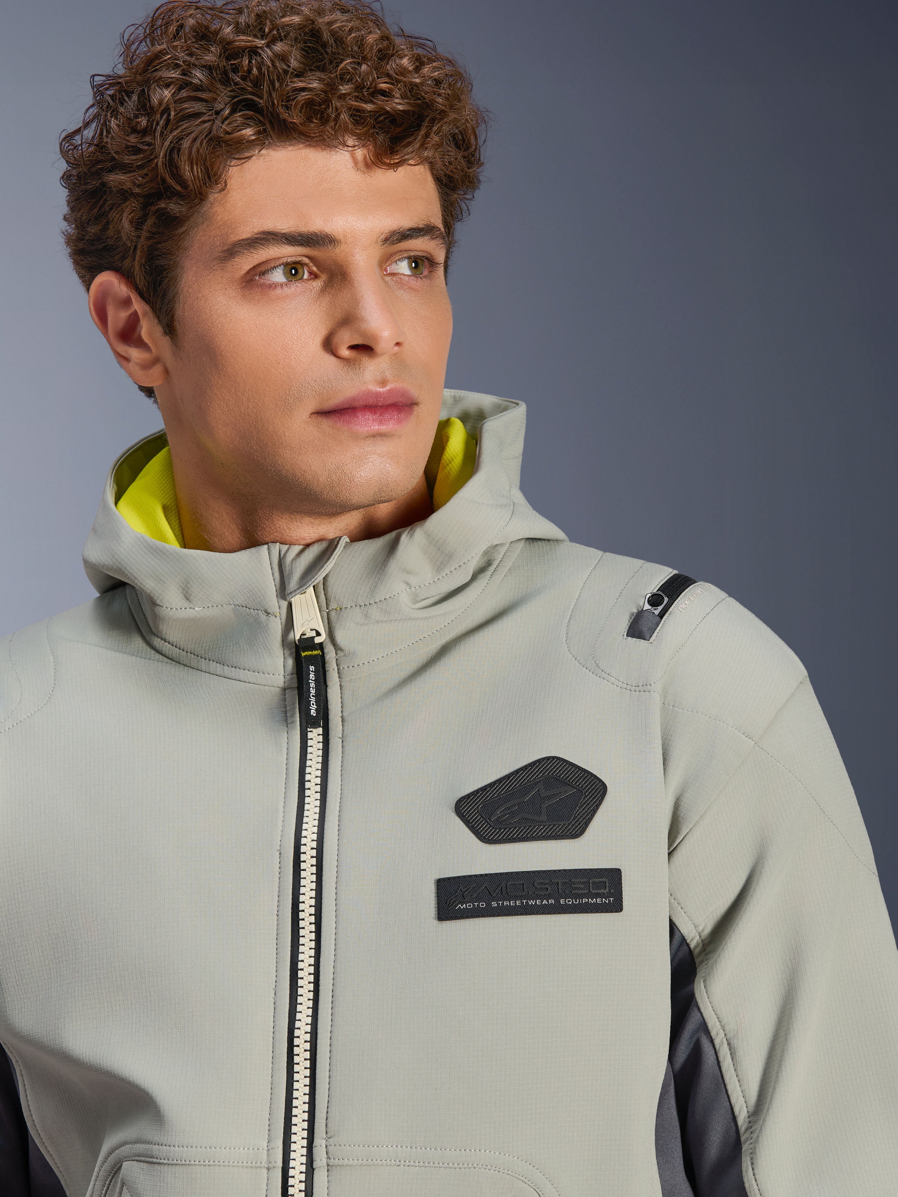Chaqueta impermeable Alpinestars Moblast, Iron Gray Dark Sage, primer plano de un modelo masculino con la chaqueta, con el logotipo de Alpinestars en negro y parches MO.ST.EQ. en el pecho, forro de la capucha en amarillo brillante y un diseño técnico moderno.