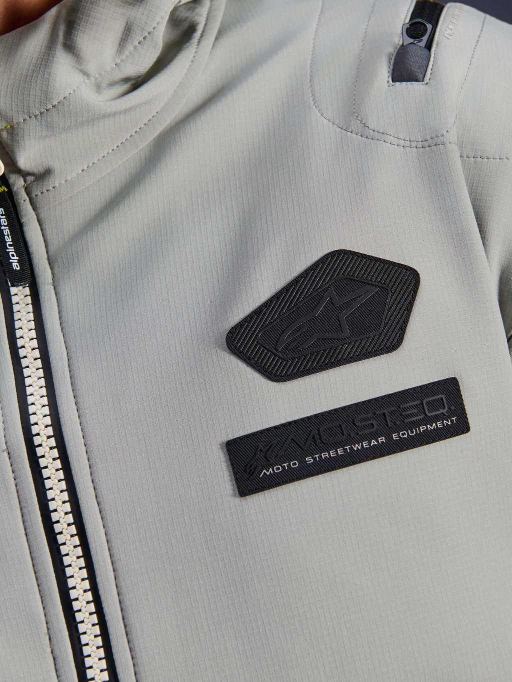 Chaqueta impermeable Alpinestars Moblast, gris hierro, detalle de cerca de los parches del pecho con el logotipo de Alpinestars y la marca MO.ST.EQ. Moto Streetwear Equipment, tejido ripstop técnico con cremallera frontal resistente y detalle de bolsillo en el hombro.