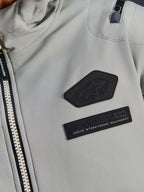 Chaqueta impermeable Alpinestars Moblast, gris hierro, detalle de cerca de los parches del pecho con el logotipo de Alpinestars y la marca MO.ST.EQ. Moto Streetwear Equipment, tejido ripstop técnico con cremallera frontal resistente y detalle de bolsillo en el hombro.