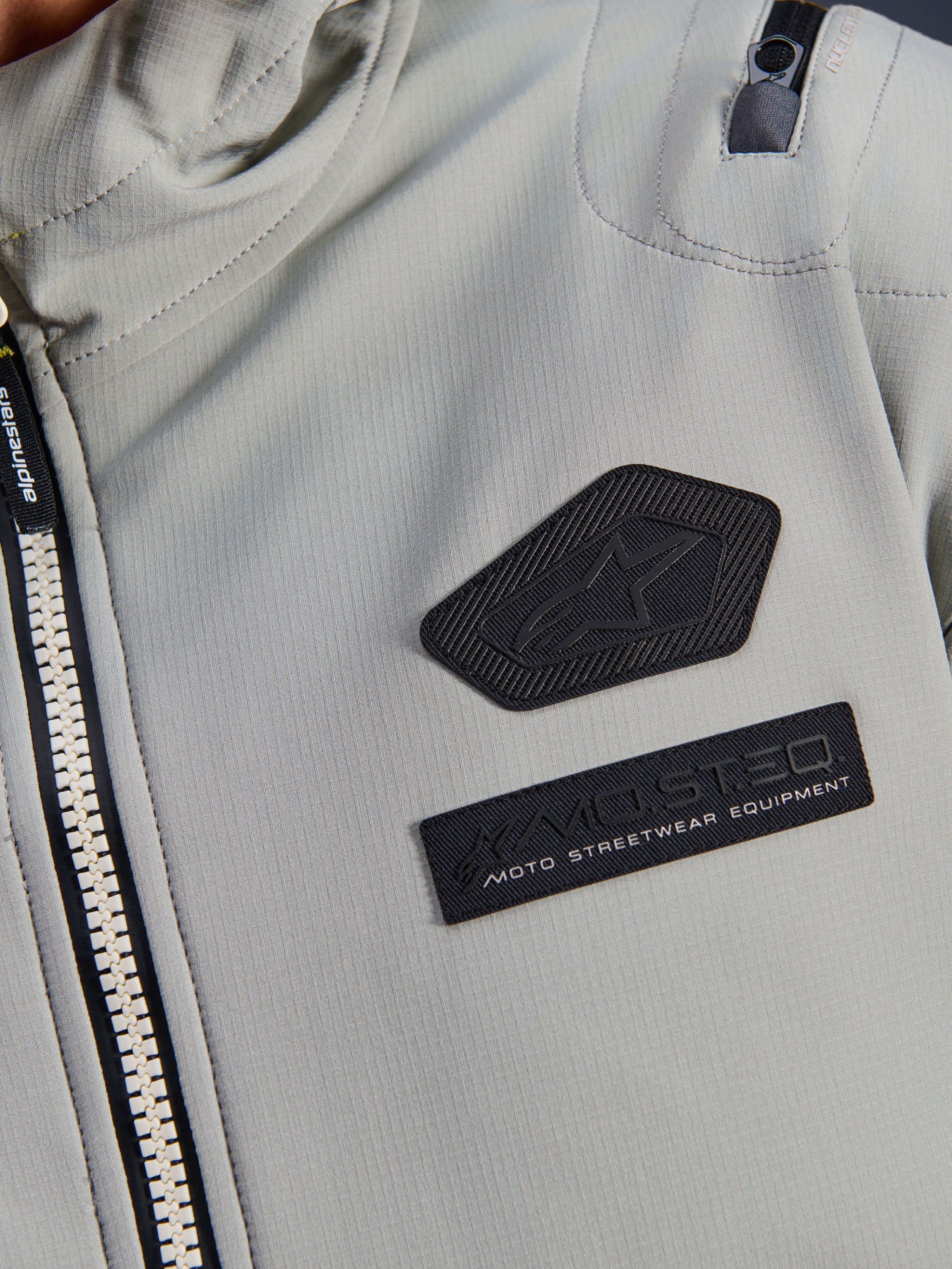Chaqueta impermeable Alpinestars Moblast, gris hierro, detalle de cerca de los parches del pecho con el logotipo de Alpinestars y la marca MO.ST.EQ. Moto Streetwear Equipment, tejido ripstop técnico con cremallera frontal resistente y detalle de bolsillo en el hombro.