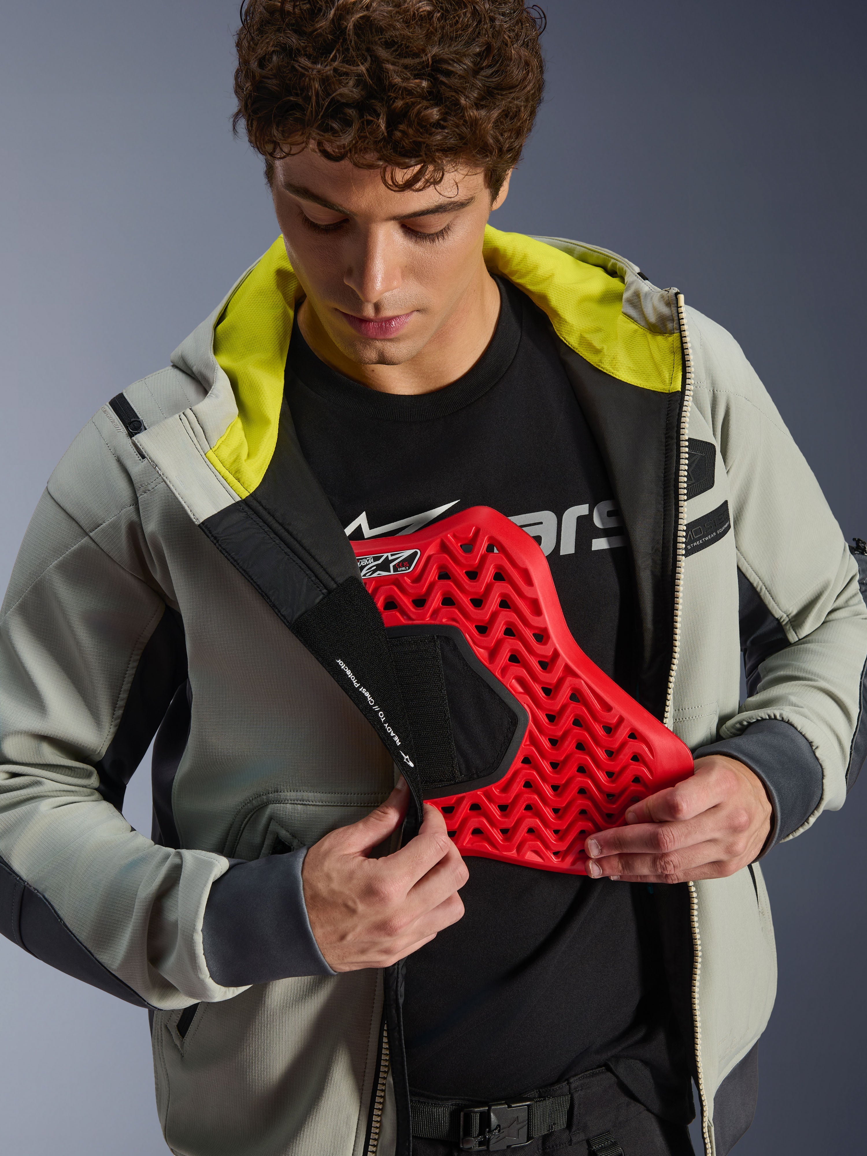 Chaqueta impermeable Alpinestars Moblast, gris hierro y verde salvia oscuro, modelo demostrando la integración de protección pectoral con un inserto rojo, forro interior verde lima brillante, equipamiento urbano moderno para motociclistas