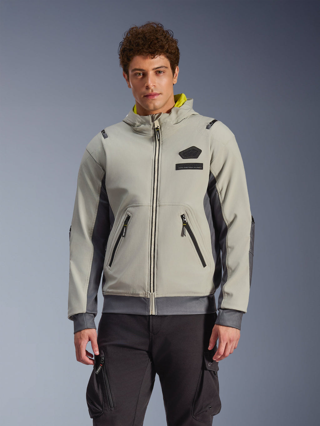 Alpinestars Moblast Waterproof Jacket, sudadera urbana para moto en gris claro y gris oscuro con forro de la capucha amarillo, bolsillos frontales y cremalleras negras, combinada con pantalones cargo negros, diseño deportivo moderno