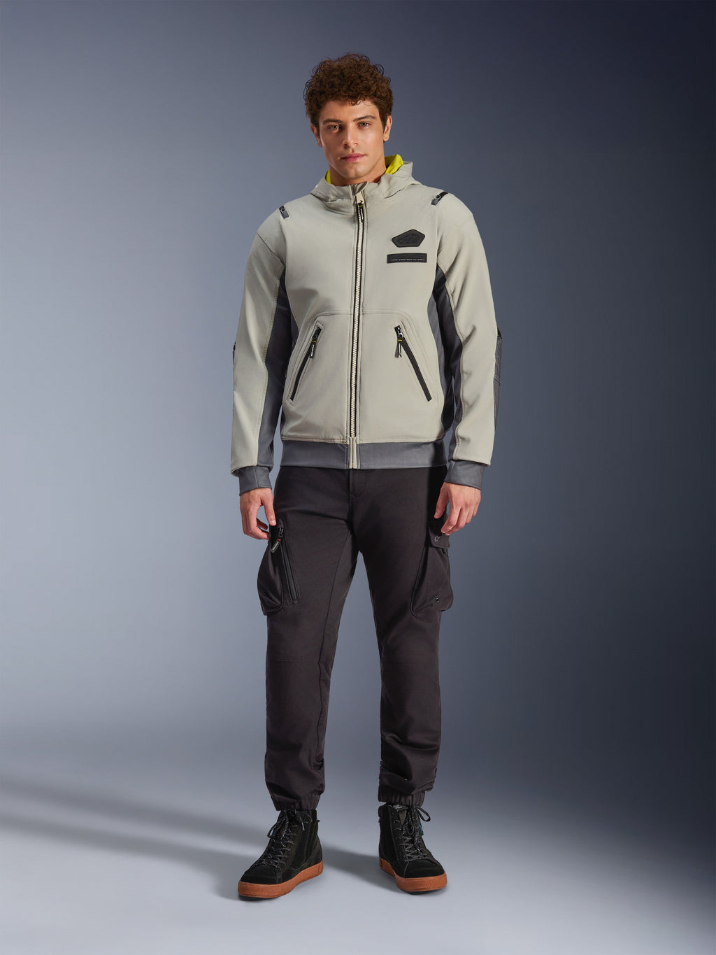 Modelo masculino vistiendo la chaqueta impermeable Alpinestars Moblast en Iron Gray y Dark Sage, chaqueta de moto con capucha de diseño técnico moderno, combinada con pantalones de montar tipo cargo oscuros y botas negras, ropa de motociclismo urbana contemporánea