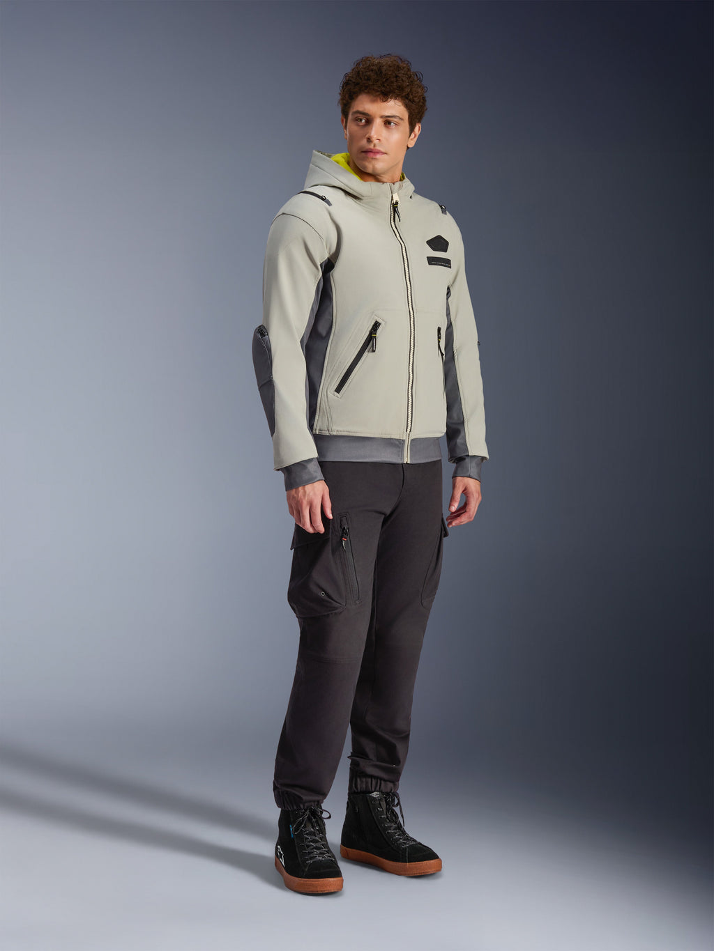 Chaqueta Alpinestars Moblast Waterproof, gris hierro y verde salvia oscuro, chaqueta de moto urbana con diseño de sudadera con capucha, usada por un modelo con pantalones de moto oscuros y botas negras, estética ciber-tecnológica minimalista con interior de capucha amarillo y cremalleras impermeables