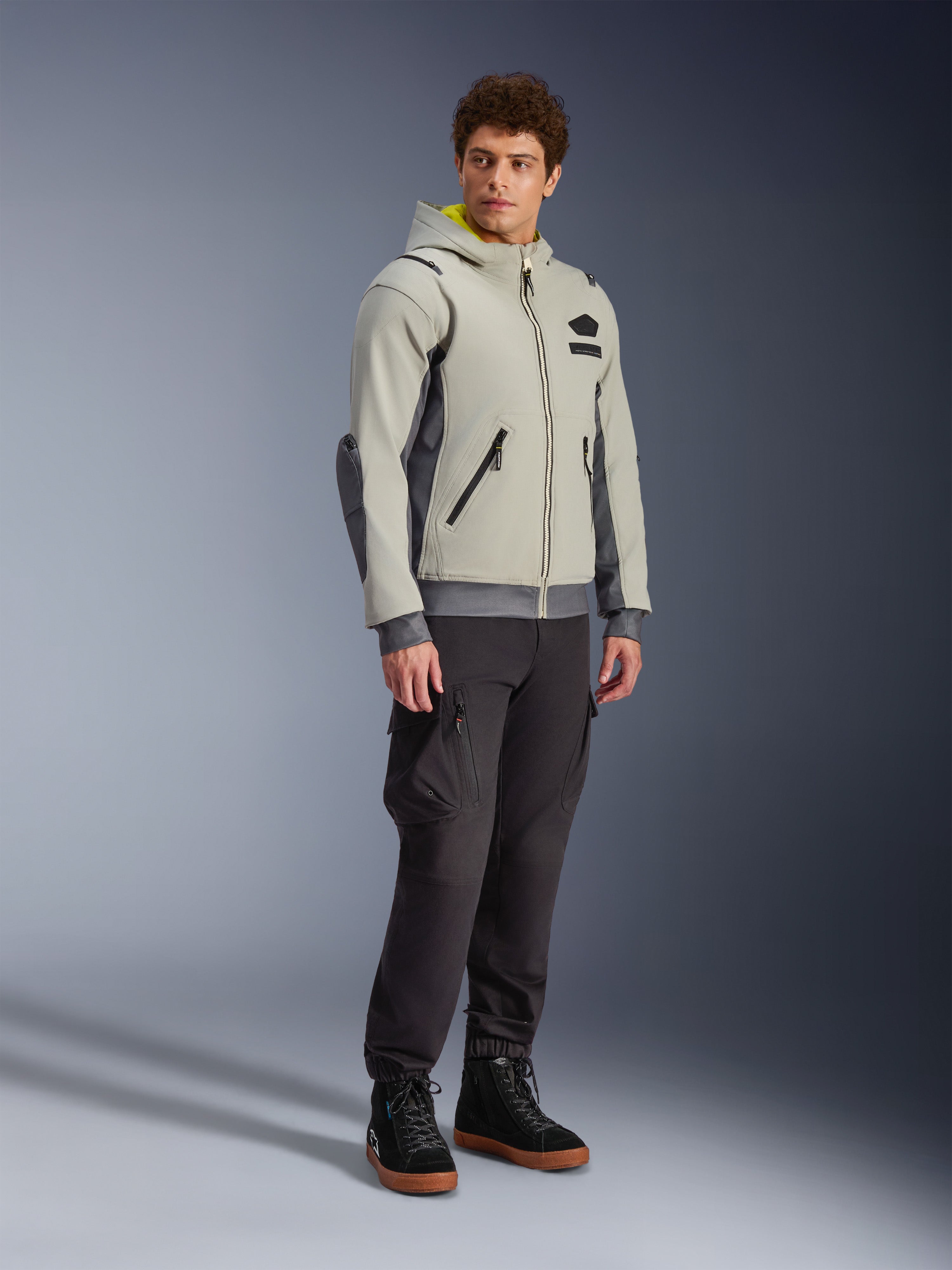 Chaqueta Alpinestars Moblast Waterproof, gris hierro y verde salvia oscuro, chaqueta de moto urbana con diseño de sudadera con capucha, usada por un modelo con pantalones de moto oscuros y botas negras, estética ciber-tecnológica minimalista con interior de capucha amarillo y cremalleras impermeables