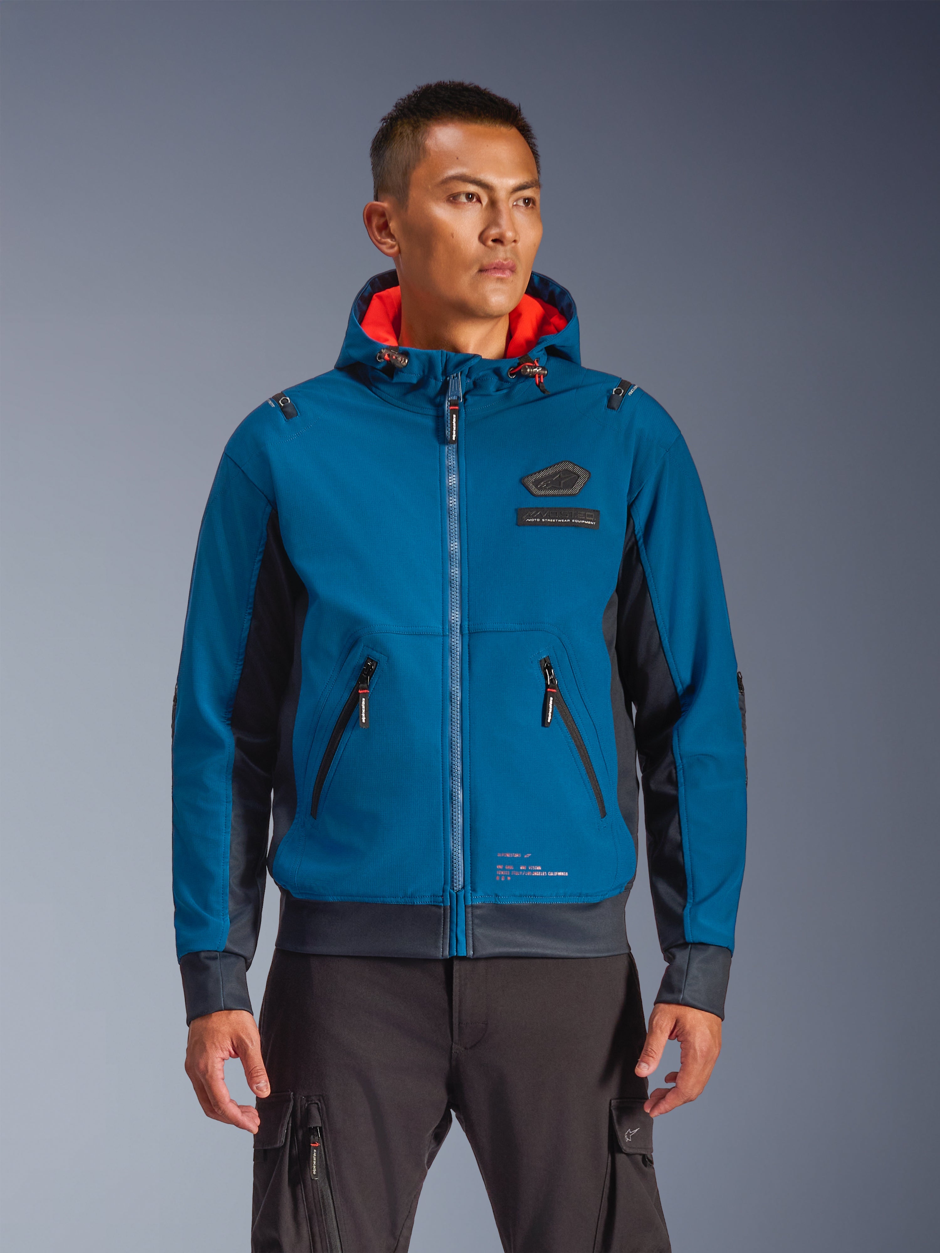Alpinestars Moblast Waterproof Jacket usada por un modelo, chaqueta de moto para conducción urbana en azul y azul marino, diseño con capucha y forro interior rojo, cremalleras negras y detalle de parche en el pecho, equipamiento de moto inspirado en el streetwear