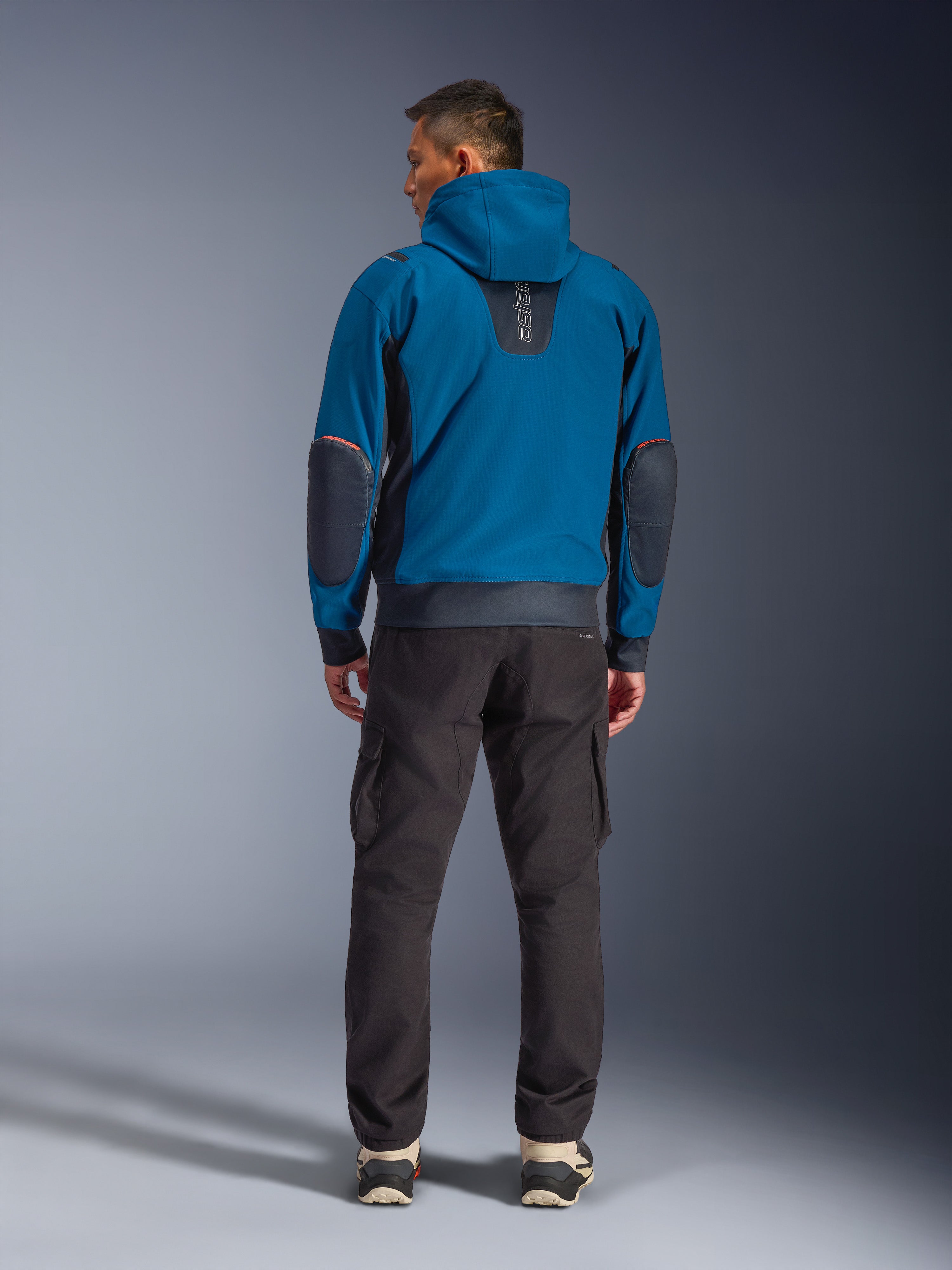 Chaqueta impermeable Alpinestars Moblast, chaqueta de moto urbana, azul petróleo oscuro y azul marino oscuro, vista trasera mostrando la capucha y los parches reforzados en los codos, usada con pantalones cargo oscuros, equipamiento elegante para trayectos diarios en cualquier clima