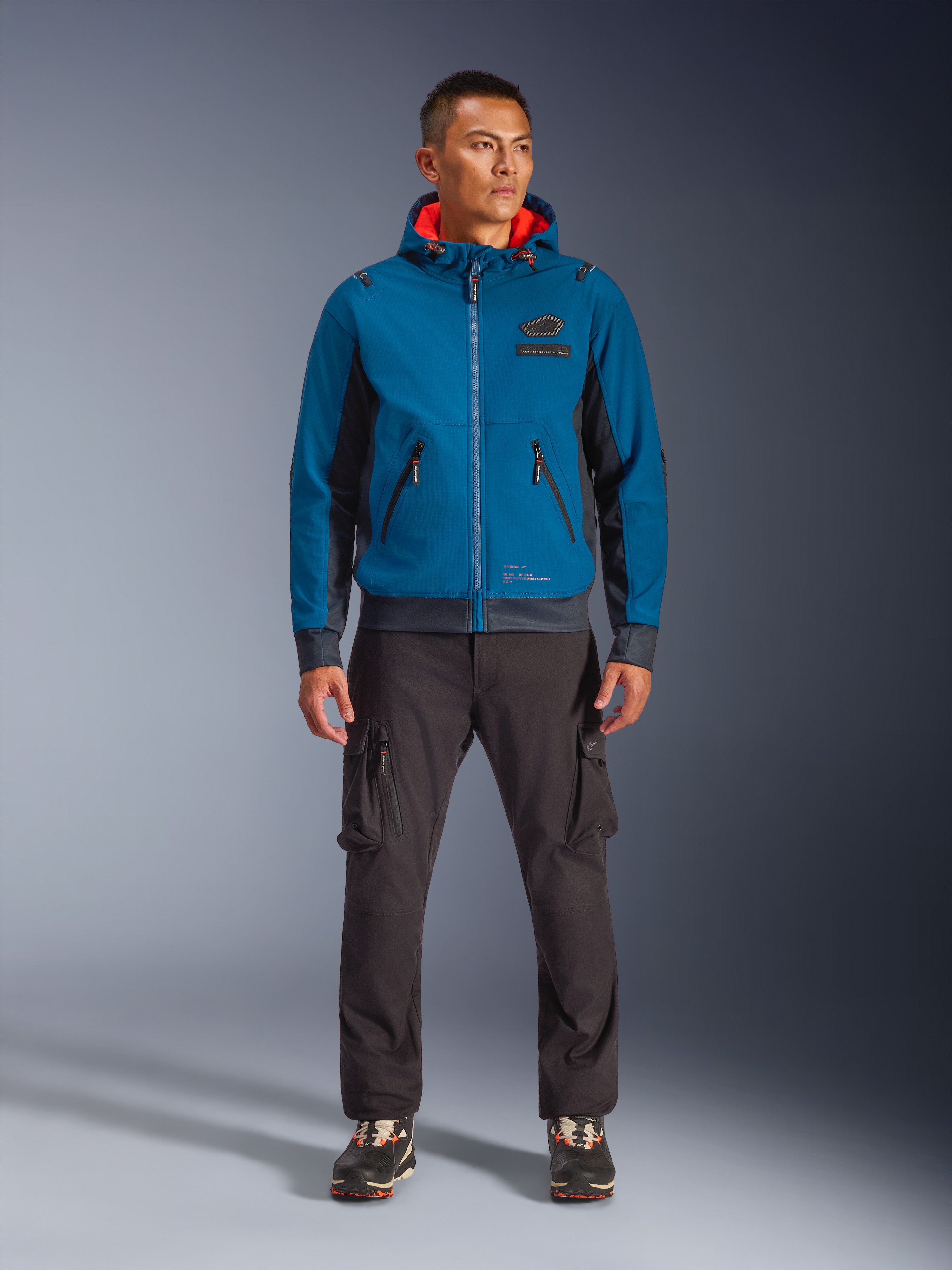 Chaqueta impermeable Alpinestars Moblast, chaqueta de moto con capucha en azul petróleo y azul marino oscuro con forro interior naranja, usada por un modelo masculino con pantalones cargo oscuros y zapatillas negras, equipamiento de conducción urbana moderno