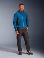 Modelo masculino vistiendo la chaqueta impermeable Alpinestars Moblast en Dark Petrol y Dark Navy, con un diseño con capucha e interior naranja brillante, combinada con pantalones cargo negros y zapatillas, equipamiento de estilo urbano moderno para motociclista.