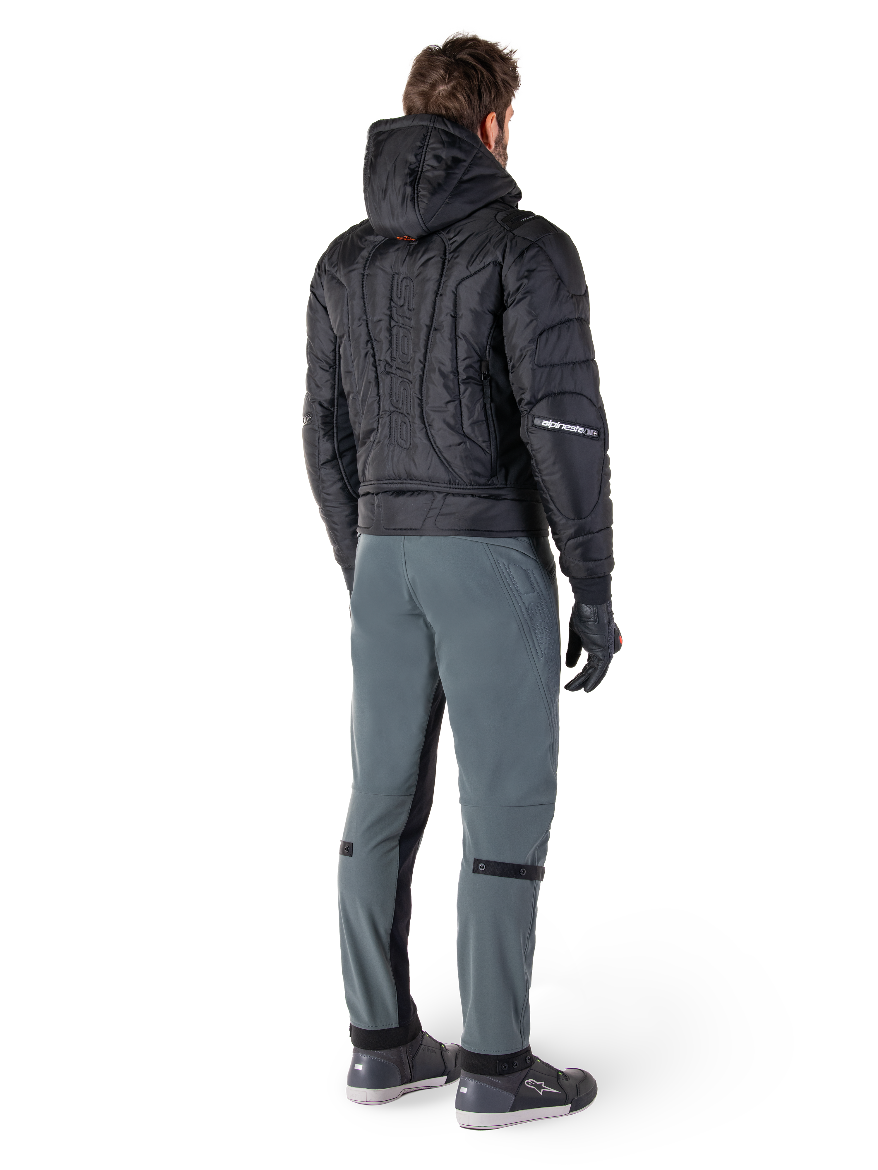 Chaqueta impermeable Alpinestars Mohobbs, negra, vista trasera de chaqueta bomber acolchada con logotipo vertical en relieve en la espalda, combinada con pantalones de conducción grises y guantes negros, equipamiento de motociclismo de estilo urbano