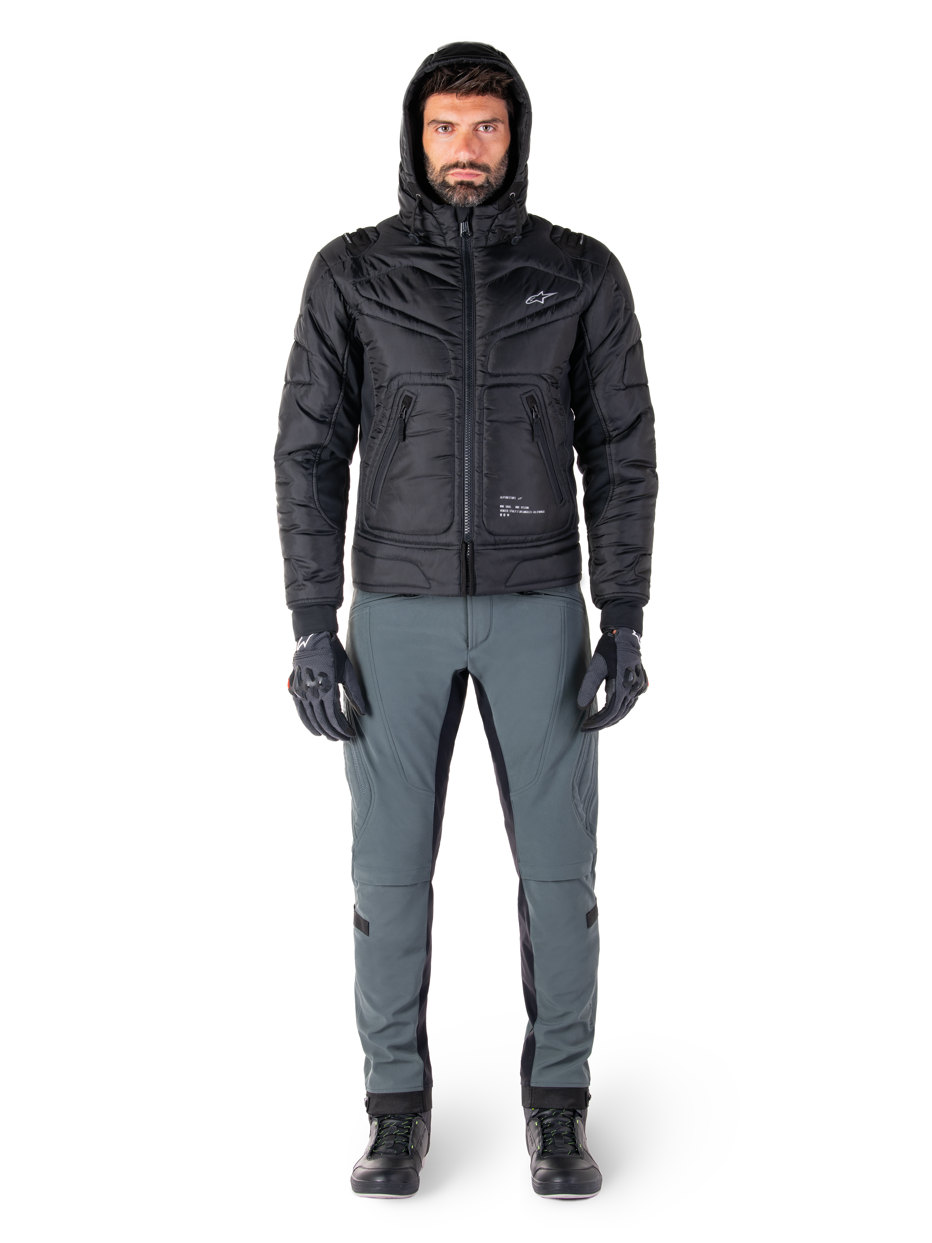 Chaqueta Alpinestars Mohobbs Waterproof en color negro, modelo masculino con chaqueta bomber acolchada y capucha, combinada con pantalones de conducción grises, guantes negros y botas negras, equipamiento de motociclismo urbano contemporáneo.