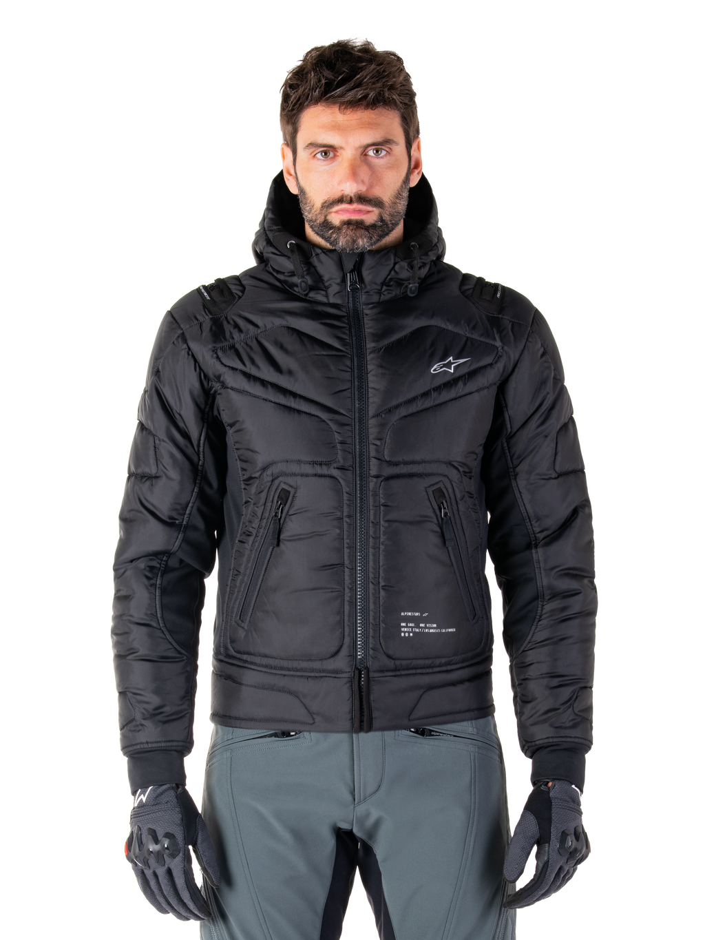 Alpinestars Mohobbs Waterproof Jacket, chaqueta bomber de moto negra acolchada, tejido de poliéster acolchado con cremallera frontal y bolsillos con cremallera, pequeño logo blanco en el pecho, usada por un modelo con pantalones grises y guantes, diseño de estilo urbano para conducción en ciudad