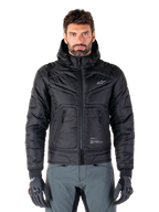 Alpinestars Mohobbs Waterproof Jacket, chaqueta bomber de moto negra acolchada, tejido de poliéster acolchado con cremallera frontal y bolsillos con cremallera, pequeño logo blanco en el pecho, usada por un modelo con pantalones grises y guantes, diseño de estilo urbano para conducción en ciudad