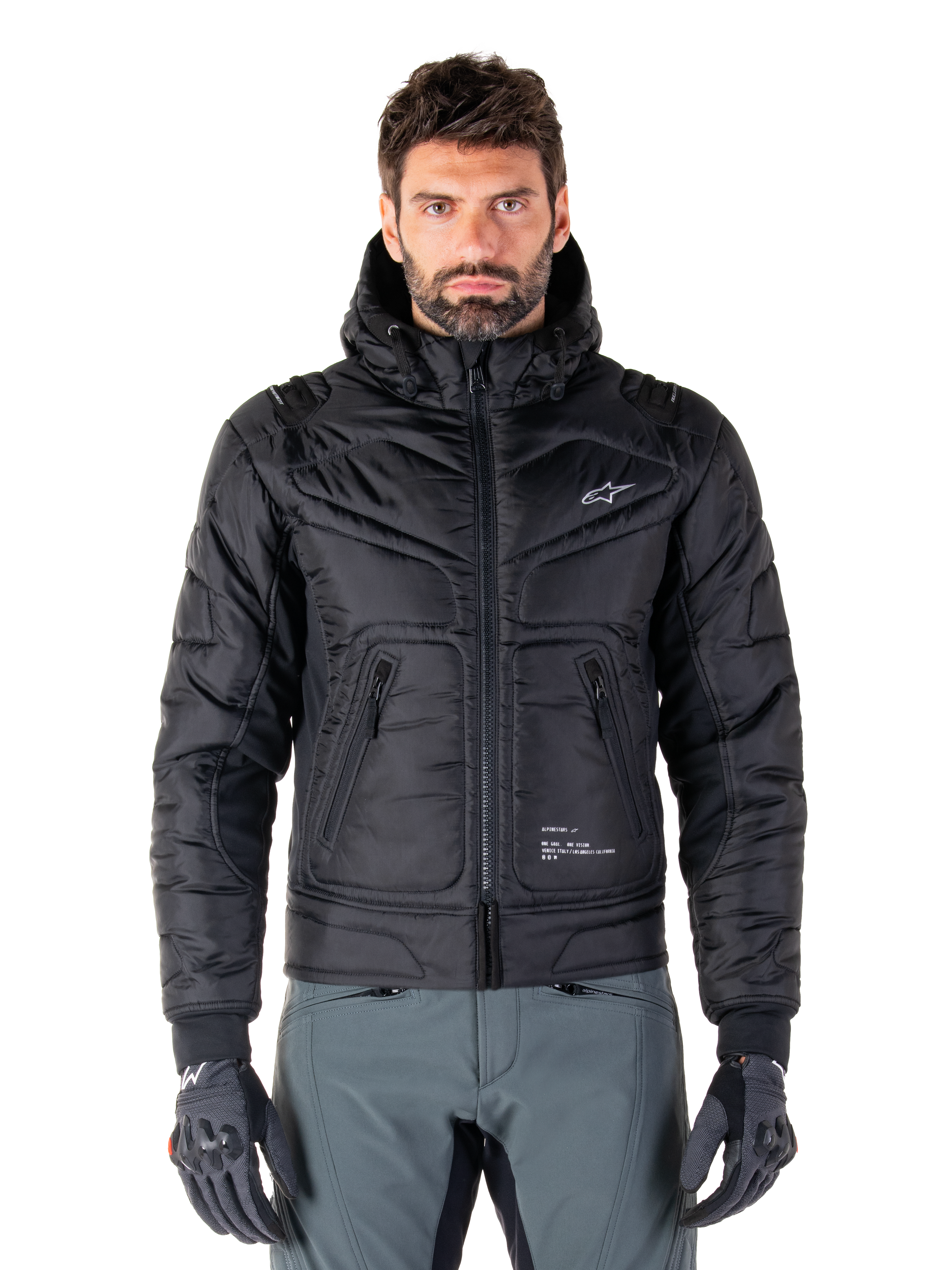 Alpinestars Mohobbs Waterproof Jacket, chaqueta bomber de moto negra acolchada, tejido de poliéster acolchado con cremallera frontal y bolsillos con cremallera, pequeño logo blanco en el pecho, usada por un modelo con pantalones grises y guantes, diseño de estilo urbano para conducción en ciudad