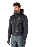 Chaqueta impermeable Mohobbs de Alpinestars, negra, diseño de chaqueta bomber acolchada con capucha, usada por un hombre con pantalones de moto grises y guantes negros, ropa de moto de inspiración urbana con un pequeño logo blanco en el pecho
