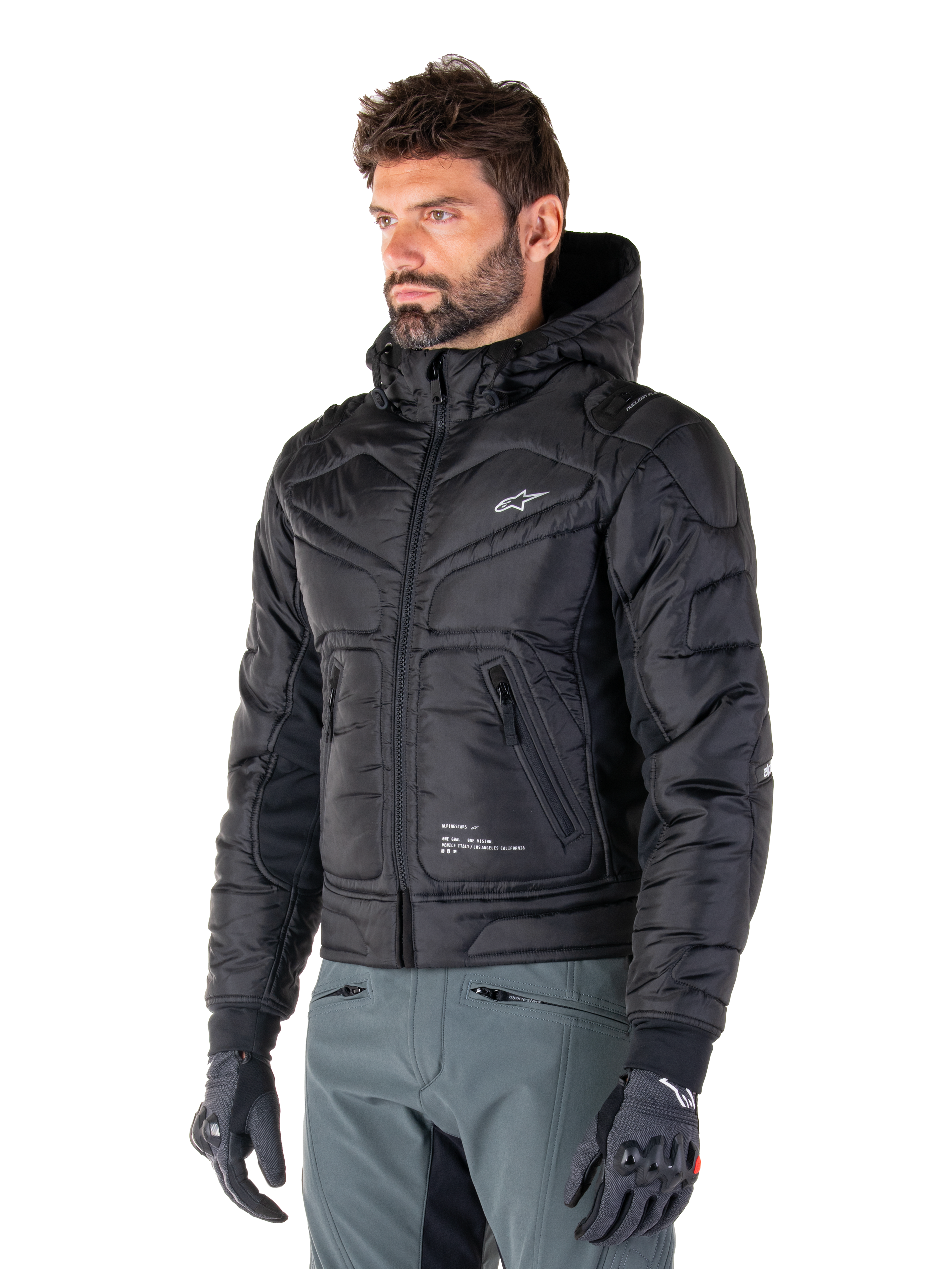 Chaqueta impermeable Mohobbs de Alpinestars, negra, diseño de chaqueta bomber acolchada con capucha, usada por un hombre con pantalones de moto grises y guantes negros, ropa de moto de inspiración urbana con un pequeño logo blanco en el pecho