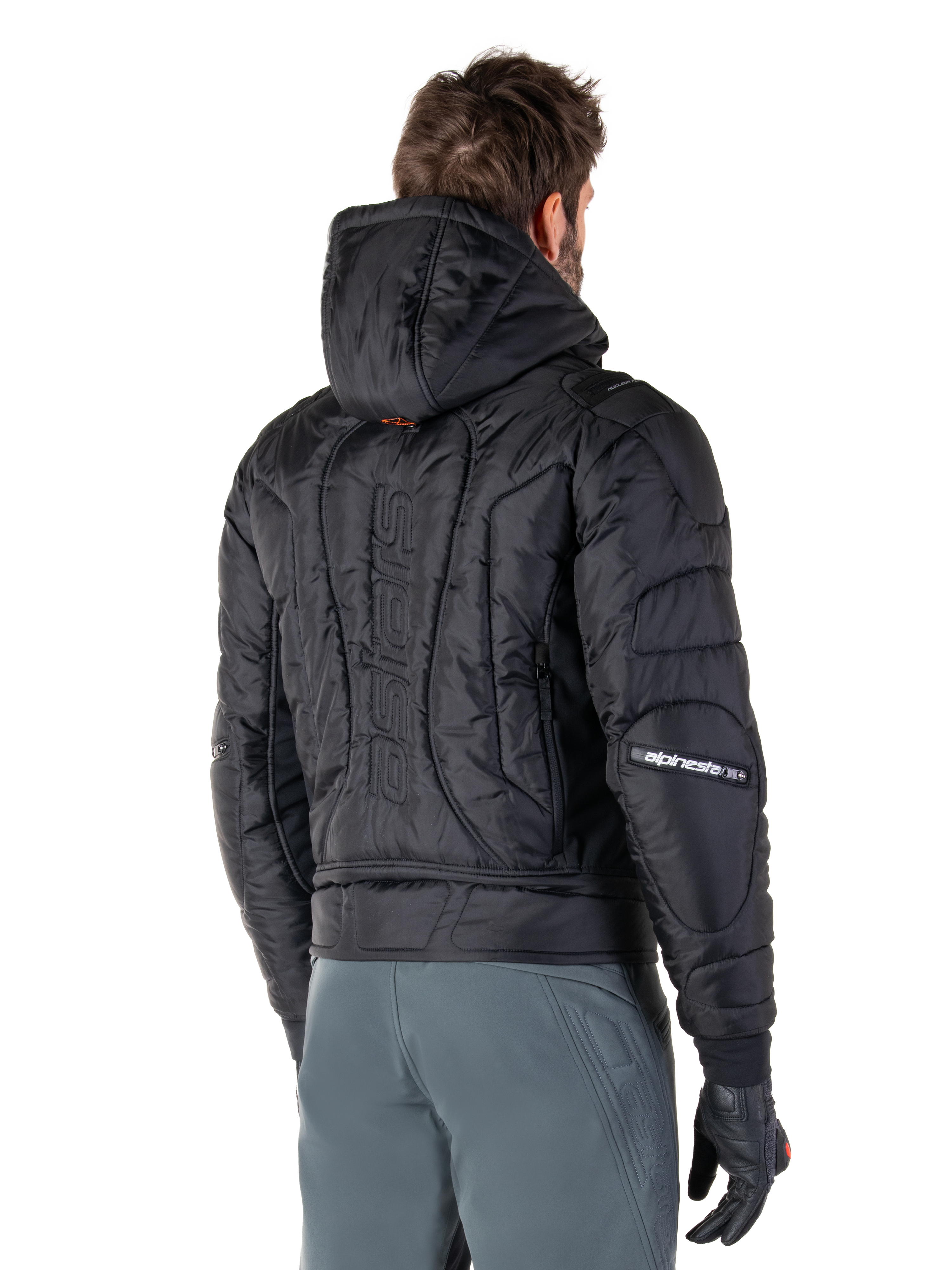 Chaqueta Alpinestars Mohobbs Waterproof, negra, vista trasera que muestra la construcción acolchada, capucha, marca denominativa 'astars' vertical en la espalda y logotipos de Alpinestars en las mangas, usada con pantalones grises y guantes negros.