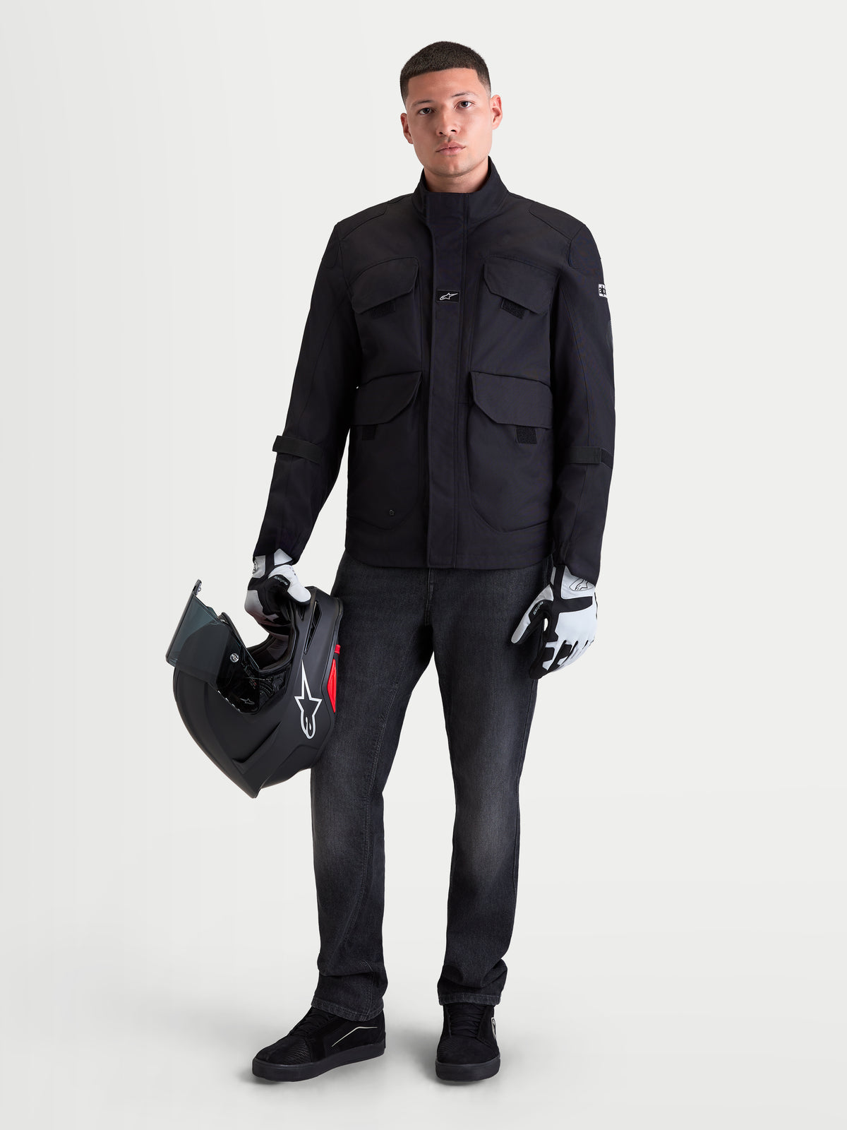 Alpinestars Altan Drystar Chaqueta, chaqueta de moto urbana contemporánea para hombre en negro, vista frontal en un modelo sosteniendo un casco y usando guantes de moto, chaqueta impermeable de tejido textil utilitario con bolsillos cargo