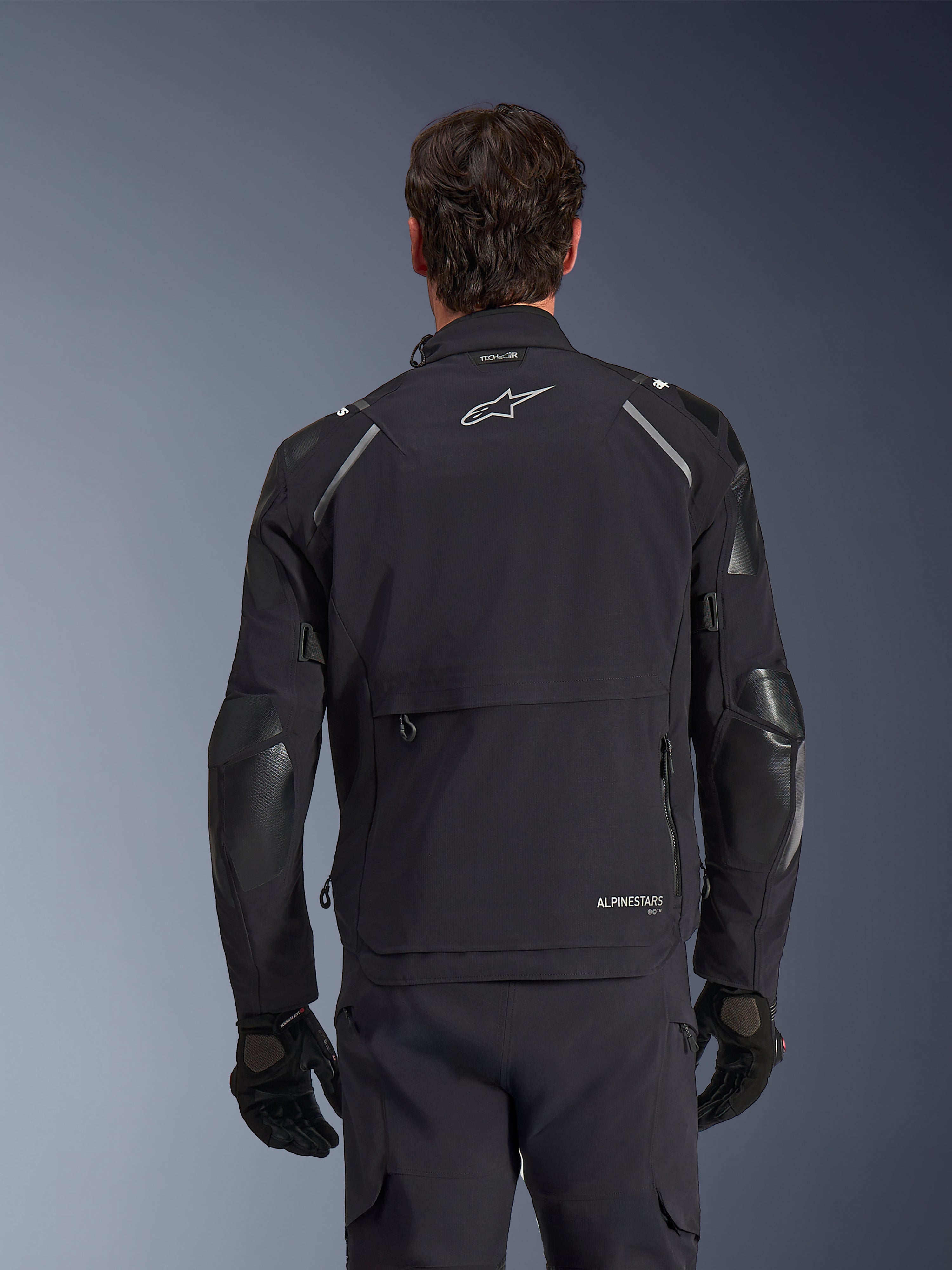Chaqueta Alpinestars Andes Pro Drystar® XF, negro y gris oscuro, vista trasera en modelo masculino, con un gran compartimento de almacenamiento trasero, detalles reflectantes en los hombros y logotipos de la marca, combinada con guantes y pantalones negros para mototurismo de aventura.