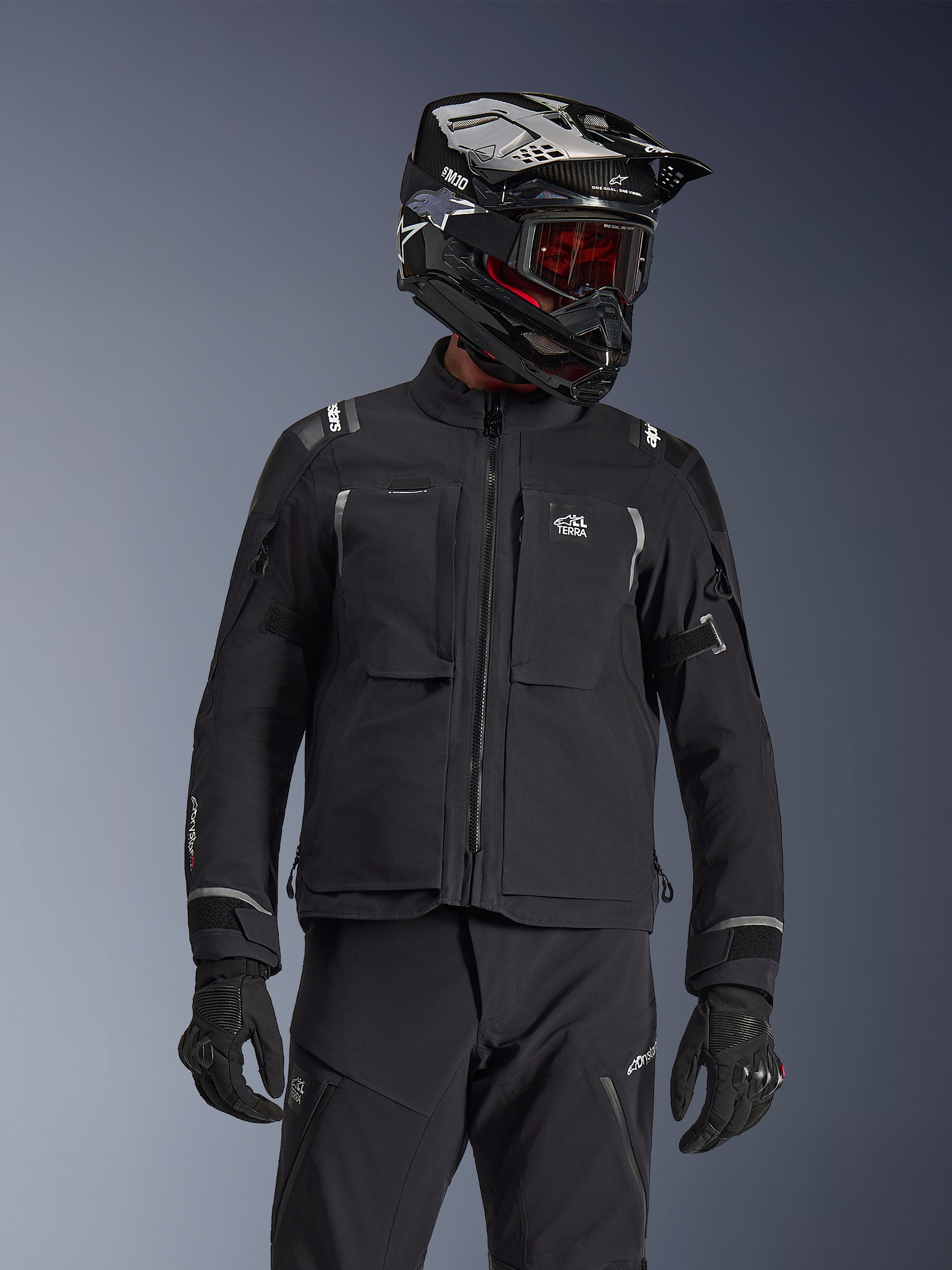 Motociclista de aventura con la chaqueta Alpinestars Andes Pro Drystar® XF en color negro y gris oscuro, con el logotipo All Terra en el pecho, combinada con pantalones técnicos a juego y un casco de aventura negro con gafas, conjunto de equipamiento profesional para touring.