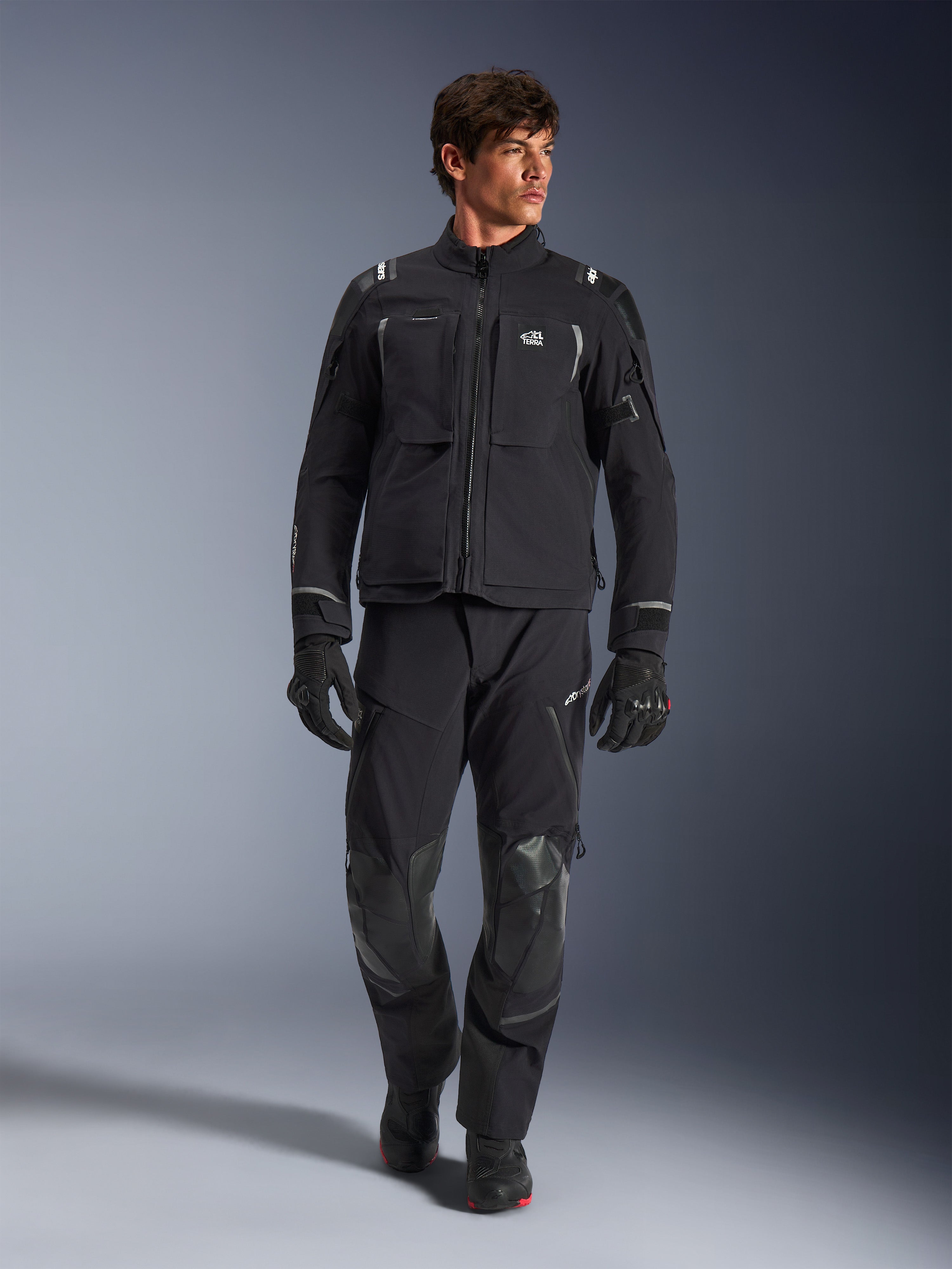 Modelo masculino con chaqueta Alpinestars Andes Pro Drystar® XF, chaqueta de moto de aventura en negro y gris oscuro, combinada con pantalones técnicos y guantes a juego, con múltiples bolsillos y logotipo 'Terra' en el pecho.