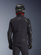 Chaqueta Alpinestars Andes Pro Drystar® XF en negro, vista trasera de un piloto con casco de fibra de carbono, con un gran compartimento de almacenamiento trasero y logotipo blanco de Alpinestars, equipamiento técnico de aventura para todas las estaciones