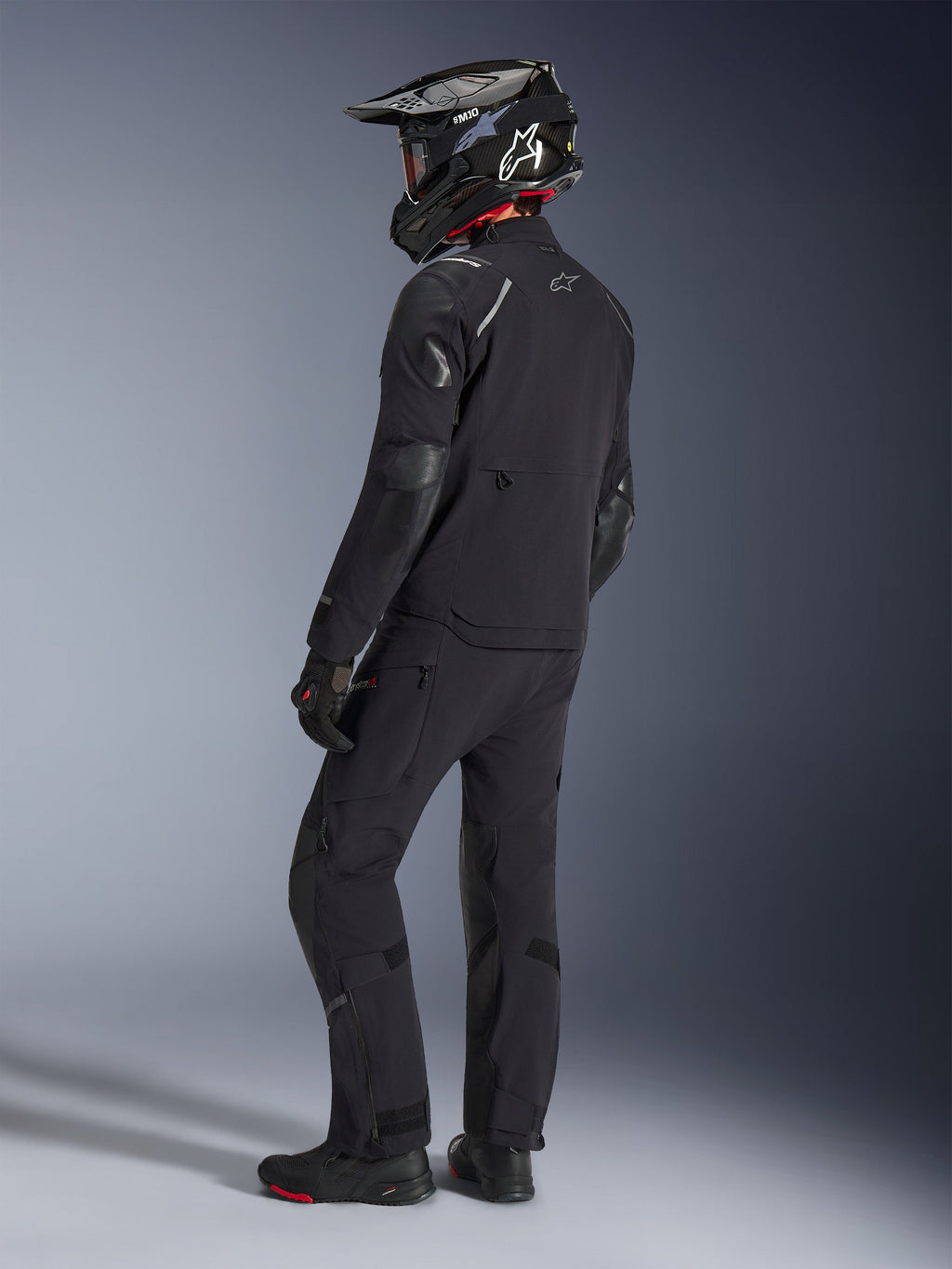 Motociclista con la chaqueta Alpinestars Andes Pro Drystar® XF y pantalones a juego en color negro y gris oscuro, vista posterior de perfil, con casco de aventura de fibra de carbono negro, guantes negros y botas de aventura, equipamiento coordinado de alto rendimiento para touring de aventura.