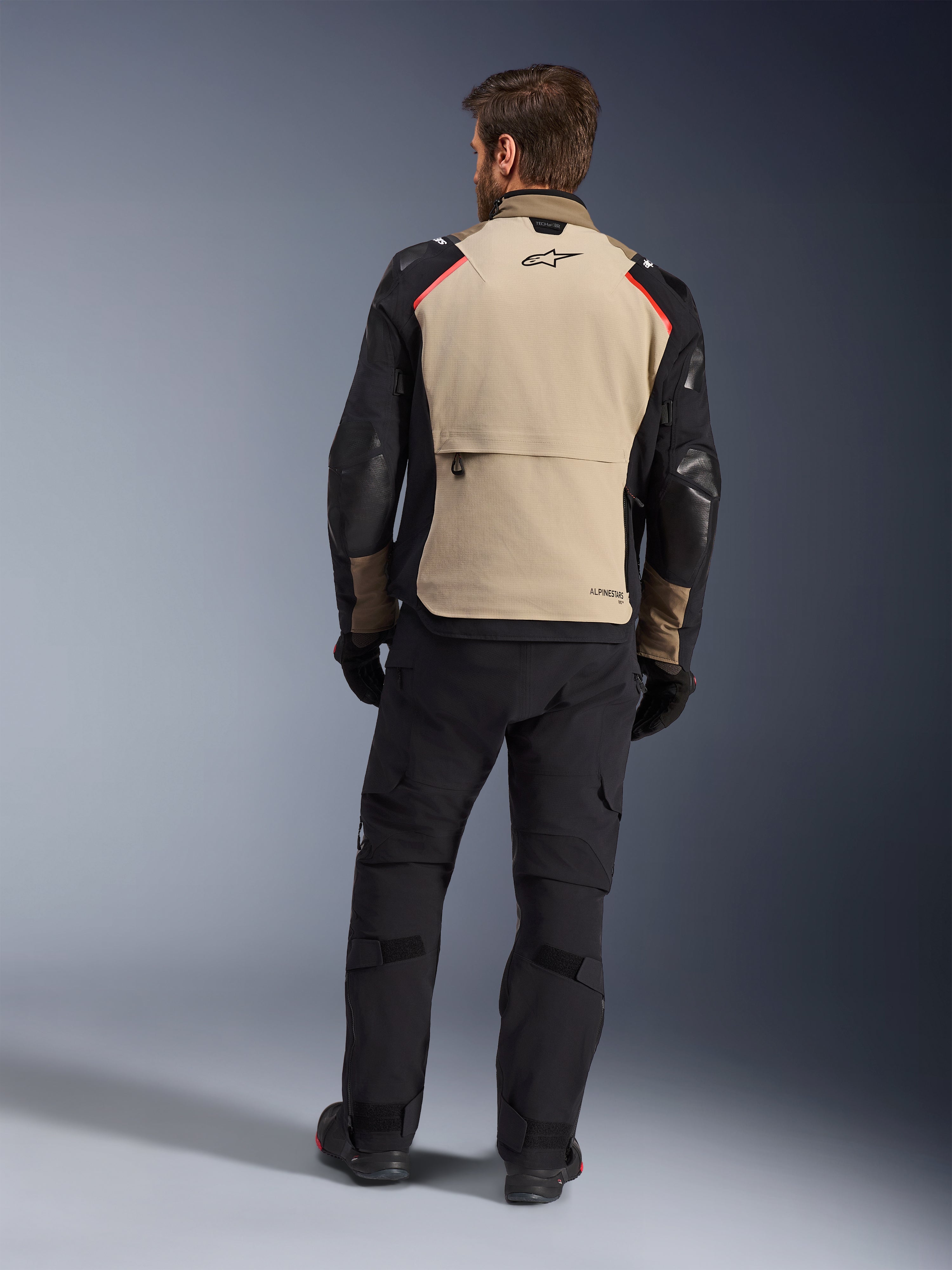 Chaqueta Alpinestars Andes Pro Drystar® XF, Walnut Aluminum, vista trasera, chaqueta de aventura para motocicleta con mangas negras y detalles rojos en los hombros, usada con pantalones de conducción y guantes negros