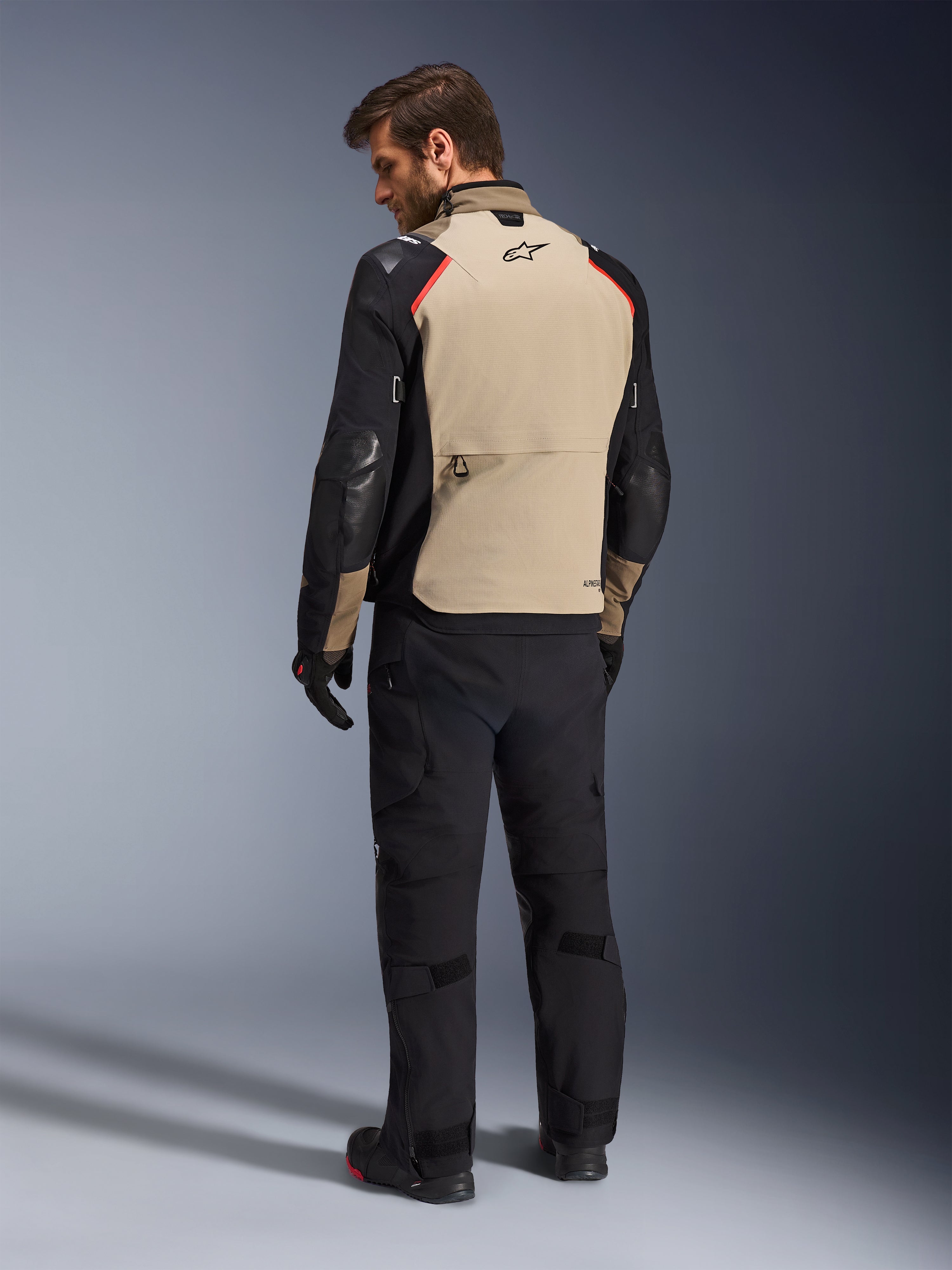 Chaqueta Alpinestars Andes Pro Drystar® XF, chaqueta de motociclismo, color nogal y negro con detalles en rojo, vista trasera en modelo, diseño de turismo de aventura con un gran bolsillo trasero, usada con pantalones de motociclismo y guantes negros.