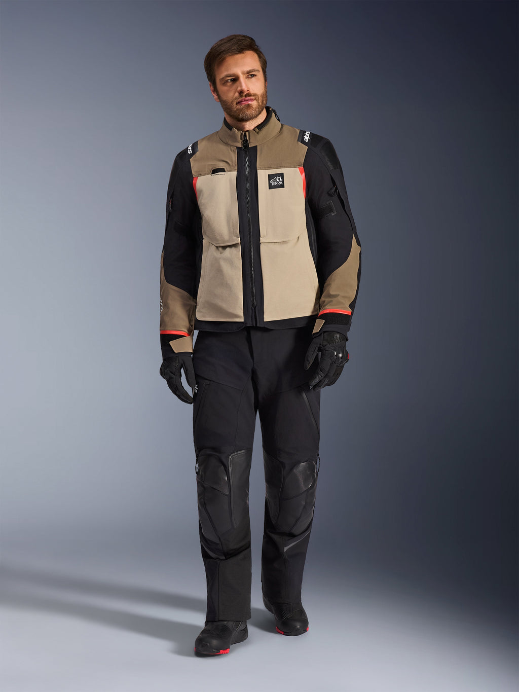 Modelo masculino vistiendo la chaqueta Alpinestars Andes Pro Drystar® XF en Walnut Aluminum, chaqueta de moto adventure touring en color canela y negro con detalles rojos, combinada con pantalones de motociclista, guantes y botas negros, toma de estudio de cuerpo completo de equipamiento de protección.