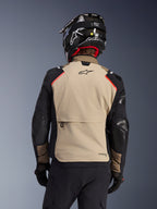 Chaqueta Alpinestars Andes Pro Drystar® XF en Walnut Aluminum, vista posterior, panel trasero de color canela con un gran bolsillo tipo cargo, mangas negras con paneles reforzados, logotipo de Alpinestars en la parte superior de la espalda, combinada con casco y guantes negros para touring de aventura.