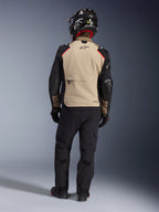 Motociclista de espaldas con la chaqueta Alpinestars Andes Pro Drystar® XF en Walnut Aluminum, panel trasero beige con mangas negras y detalles rojos, combinada con pantalones técnicos de aventura negros y casco de moto, equipamiento de touring premium para cualquier clima.