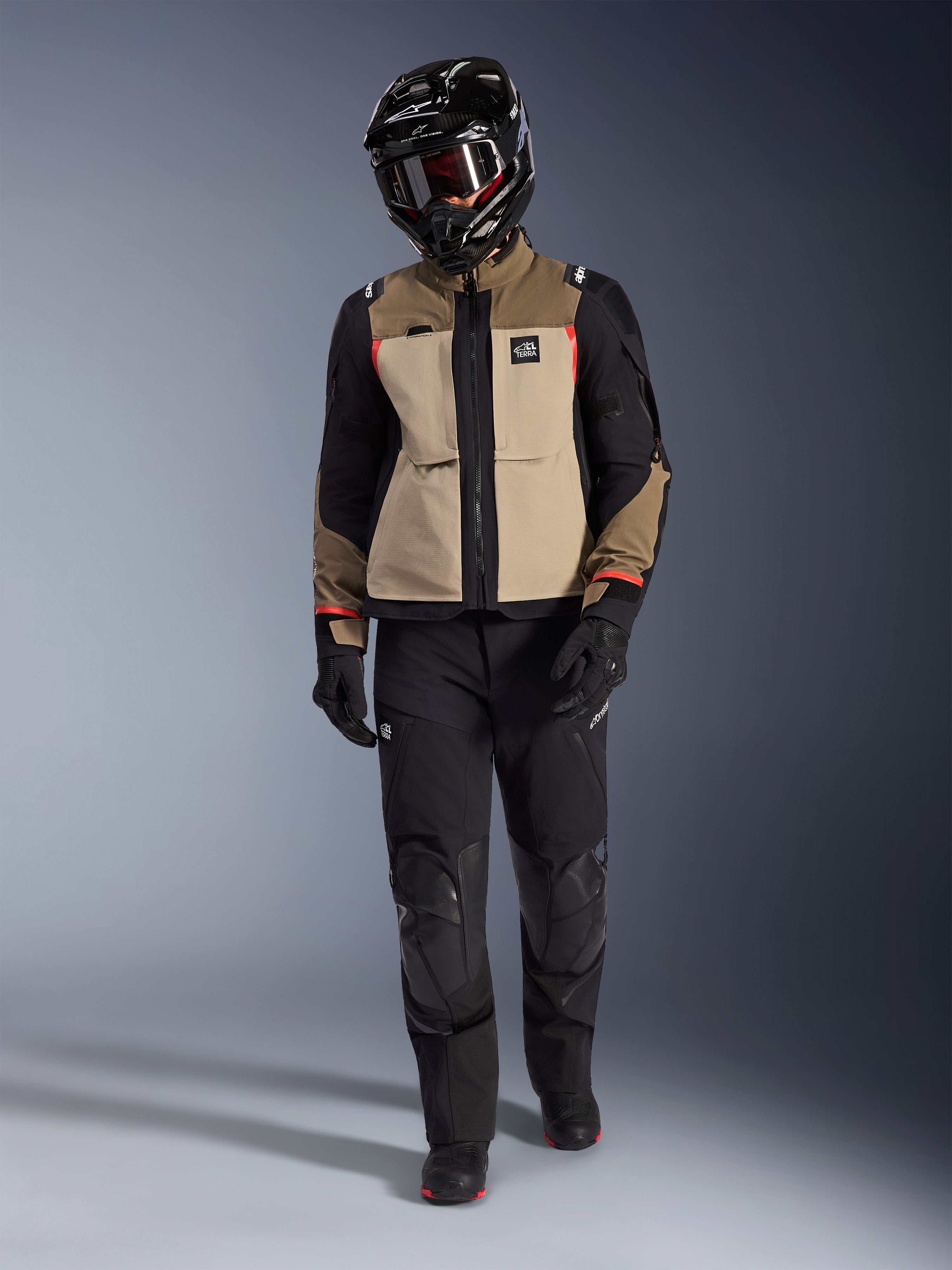 Chaqueta Alpinestars Andes Pro Drystar® XF en Walnut Aluminum, usada con pantalones de aventura negros, guantes negros y un casco integral negro con gafas, con detalles rojos y marca All Terra, equipamiento profesional para touring de aventura